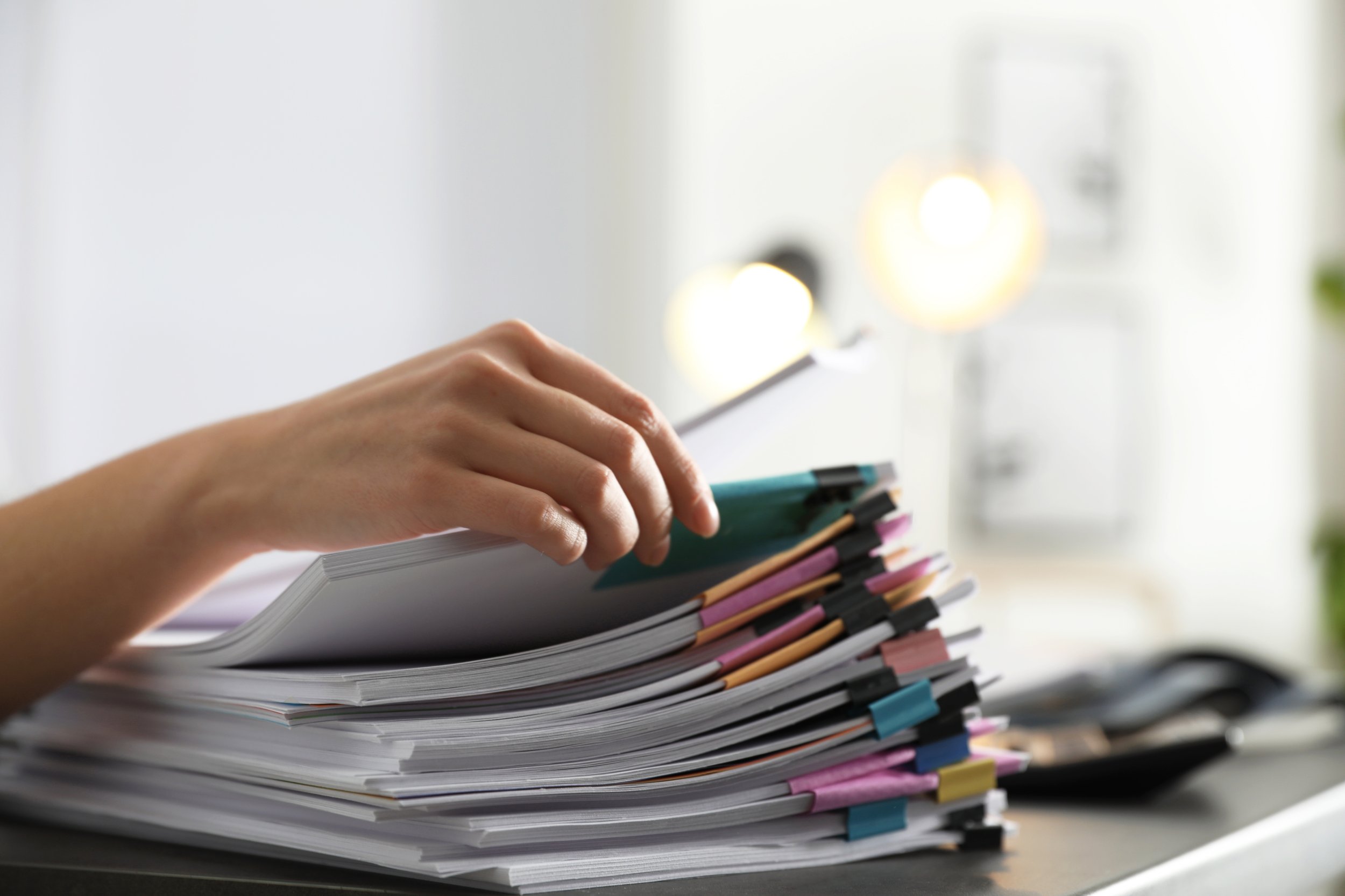 Hand reaching over a large stack of documents with colored tabs on a desk in an office setting.
