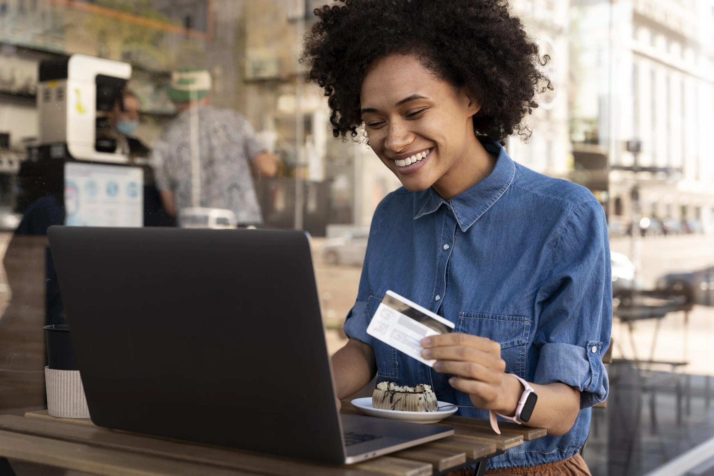 Woman shopping online at a cafe, holding a credit card, with a laptop and a cake on the table, smiling.
