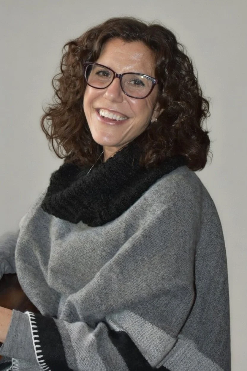 Elizabeth Kirshenbaum: A woman with curly brown hair, wearing glasses and a gray sweater with a black collar, smiling at the camera.