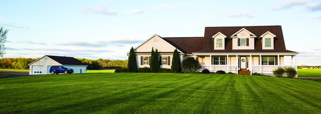 A large white house with a brown roof, surrounded by a well-manicured green lawn, and a small outbuilding with a car parked outside.
