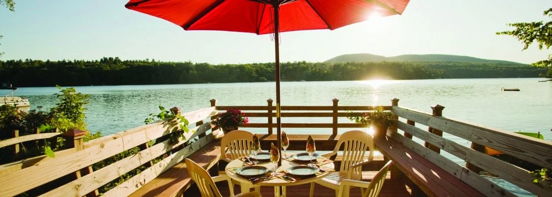 Outdoor dining table set for four with plates, wine glasses, and napkins on a deck overlooking a lake with a red umbrella providing shade.