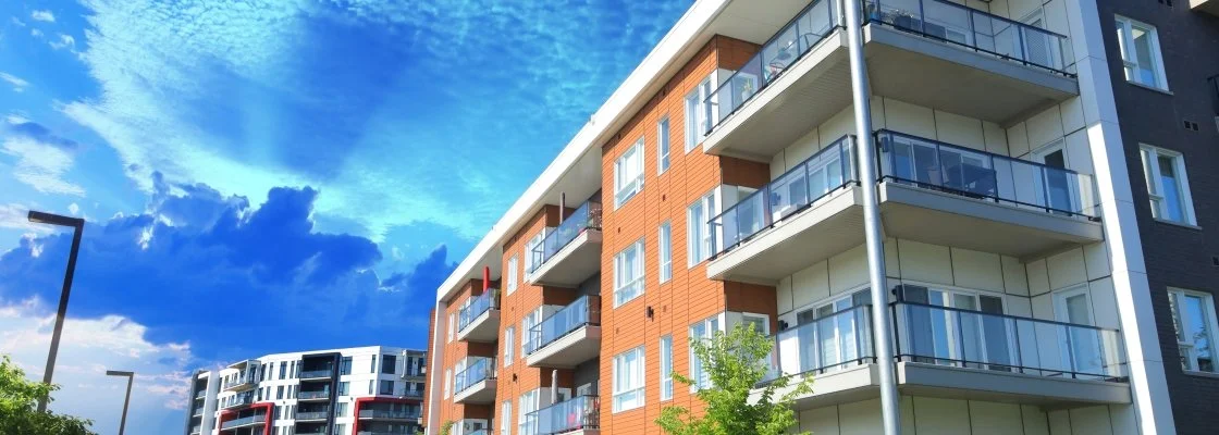 Modern multi-story apartment building with balconies, set against a bright blue sky with clouds.