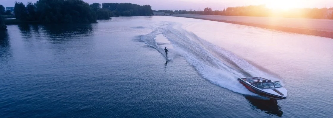 A person wakeboarding on a river during sunset, being pulled by a motorboat.