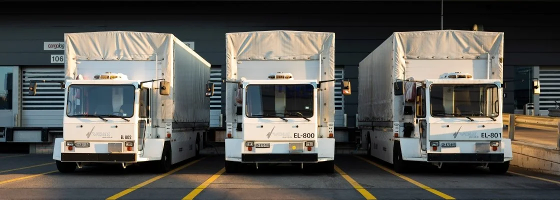 Three white delivery trucks parked in a lot in front of a warehouse.