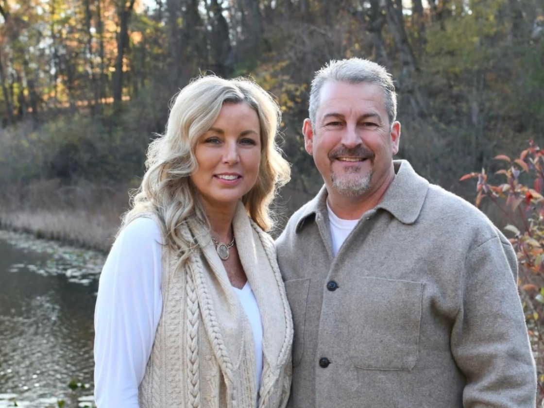 A smiling couple standing outdoors by a body of water with trees in the background, dressed in casual fall clothing.
