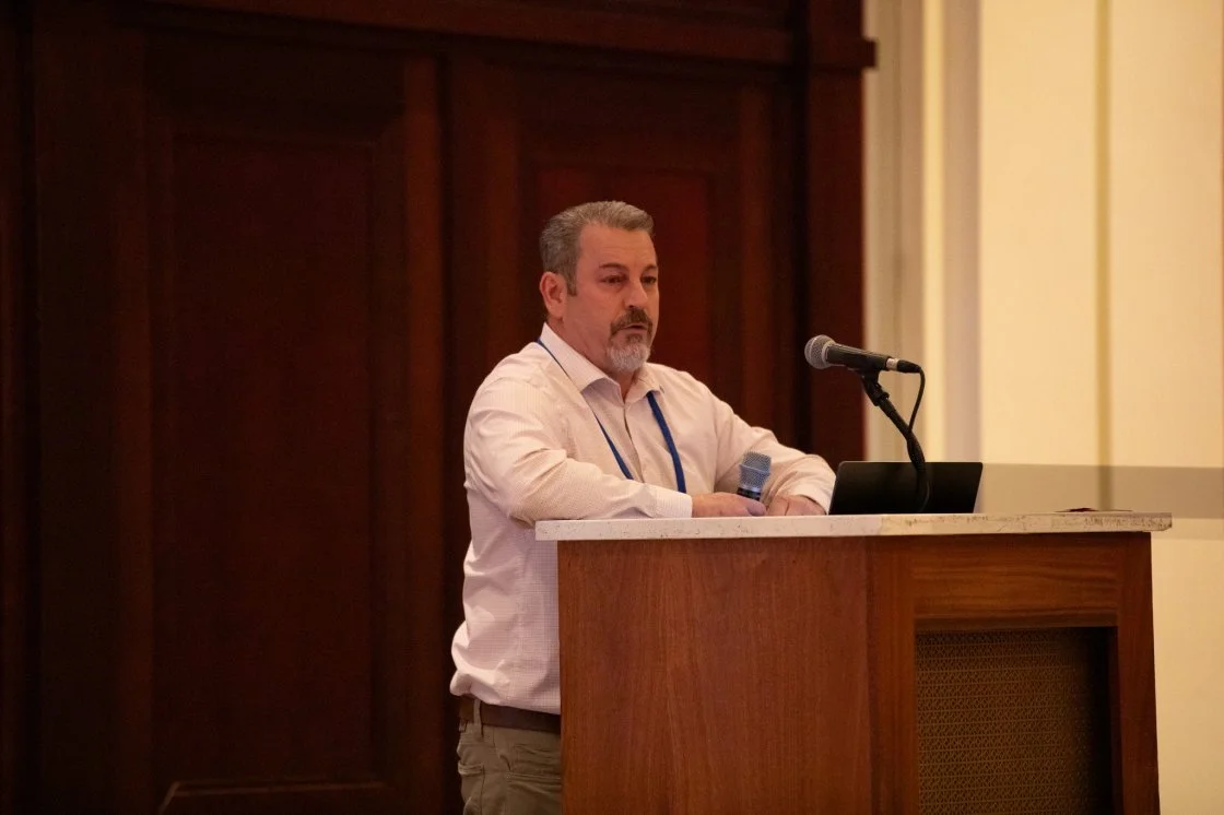 A man with a beard and short hair speaking at a podium with a microphone and a laptop in a wood-paneled room.