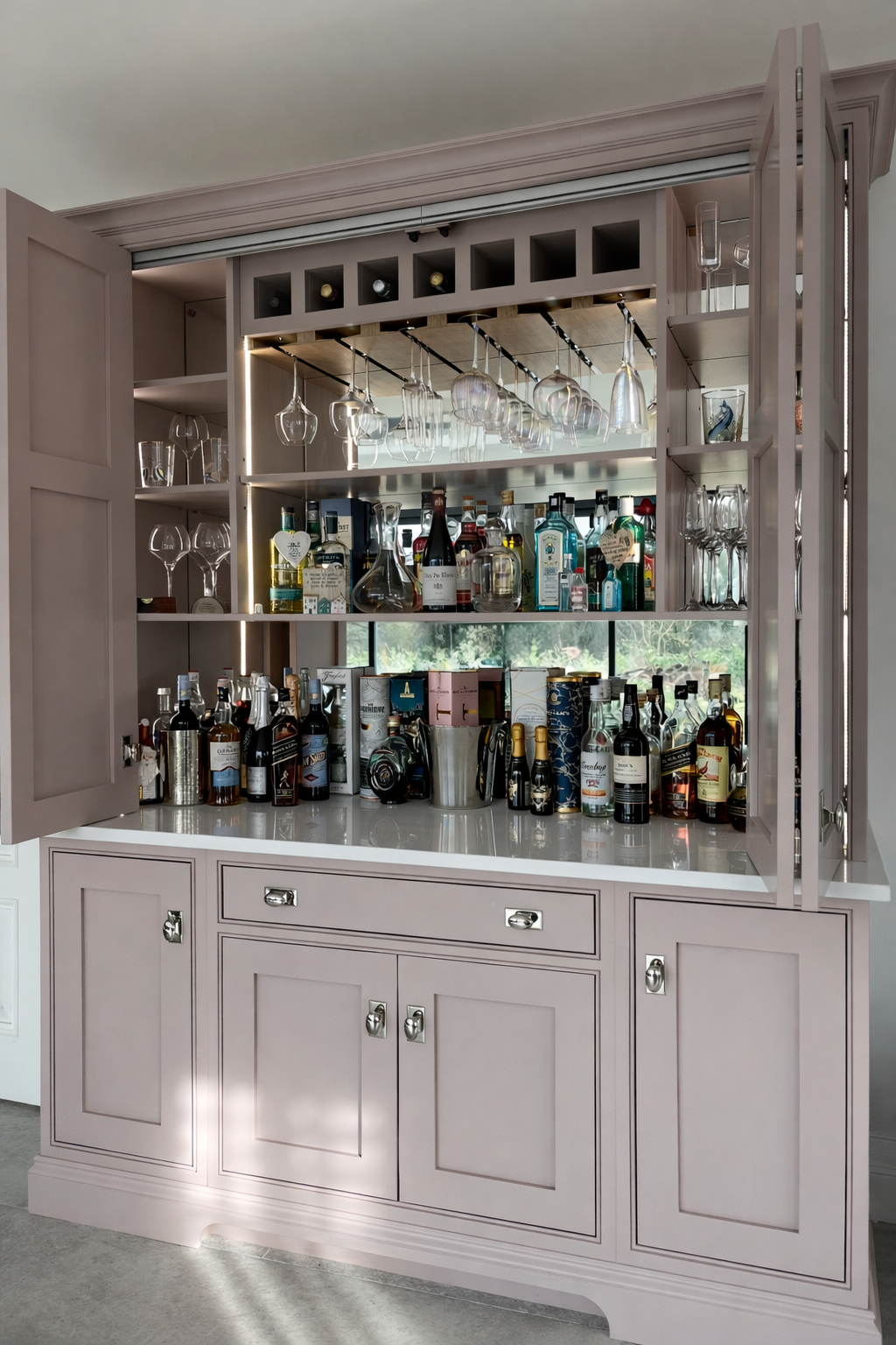 A pink bar cabinet with open doors displaying hanging wine glasses, bottles of liquor, mixers, and bar tools against a window view.