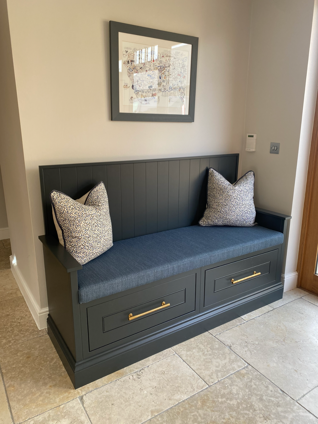 A black bench with two patterned pillows, chrome handles, and a cushioned seat, placed against a white wall with a framed map above it, next to a wooden door and a thermostat on the wall.