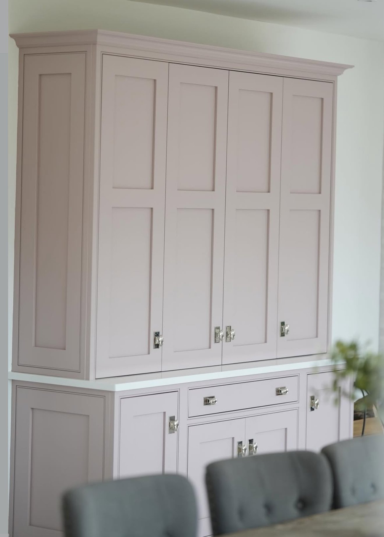 A large, light-colored wooden cabinet with multiple doors, situated against a white wall in a room with a white ceiling. Part of a dining table with a greenish-gray upholstered chair is visible in the foreground.