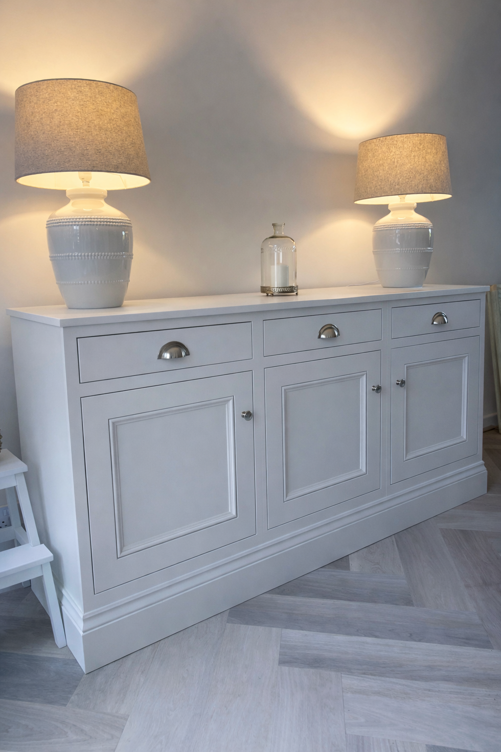 A white dresser with three drawers and two cabinet doors, topped with two matching table lamps with beige shades and a clear glass bottle in between, set against a plain light-colored wall.