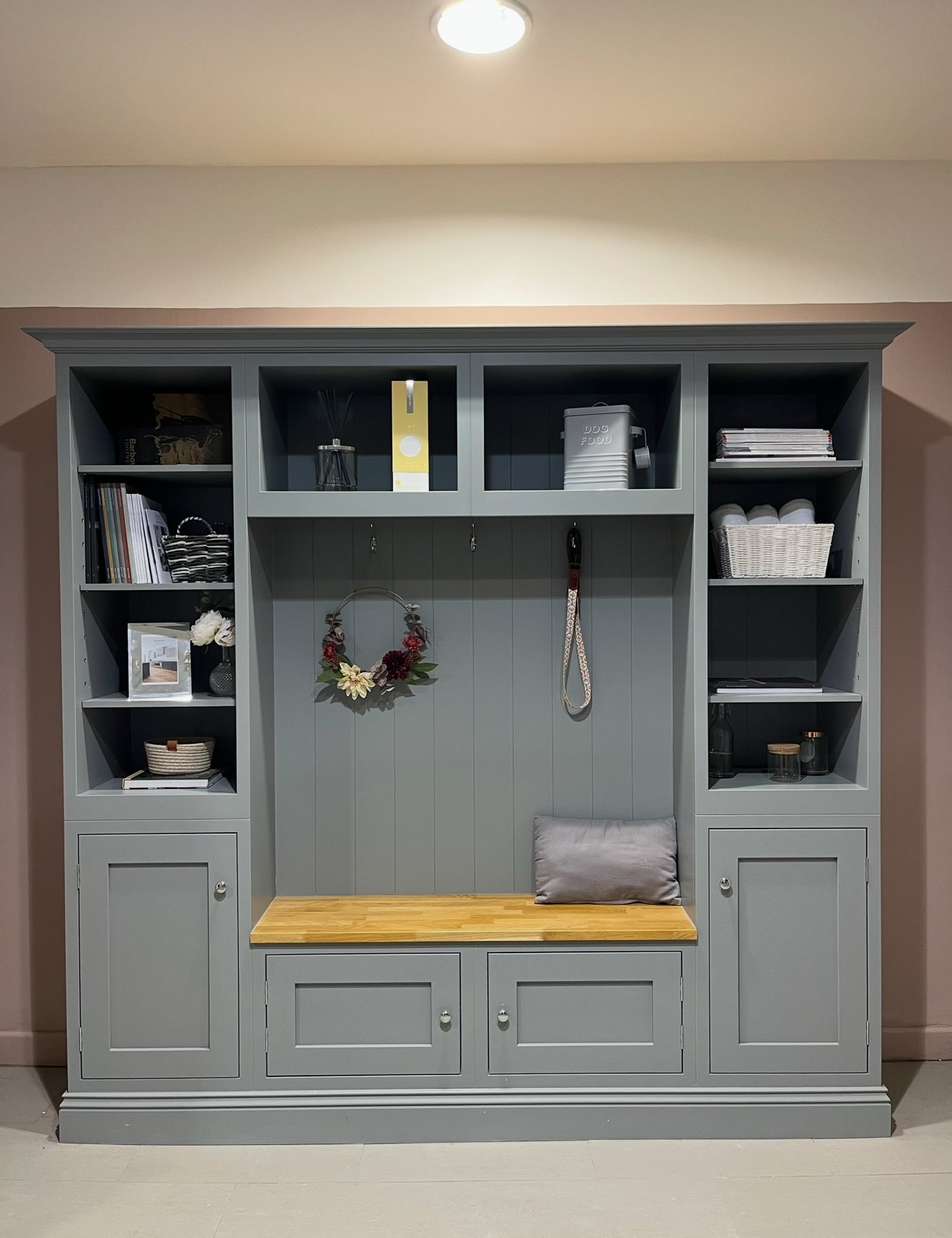 A gray built-in storage unit with open shelves and closed cabinets, featuring decorative items, books, baskets, and a wooden bench with a cushion.