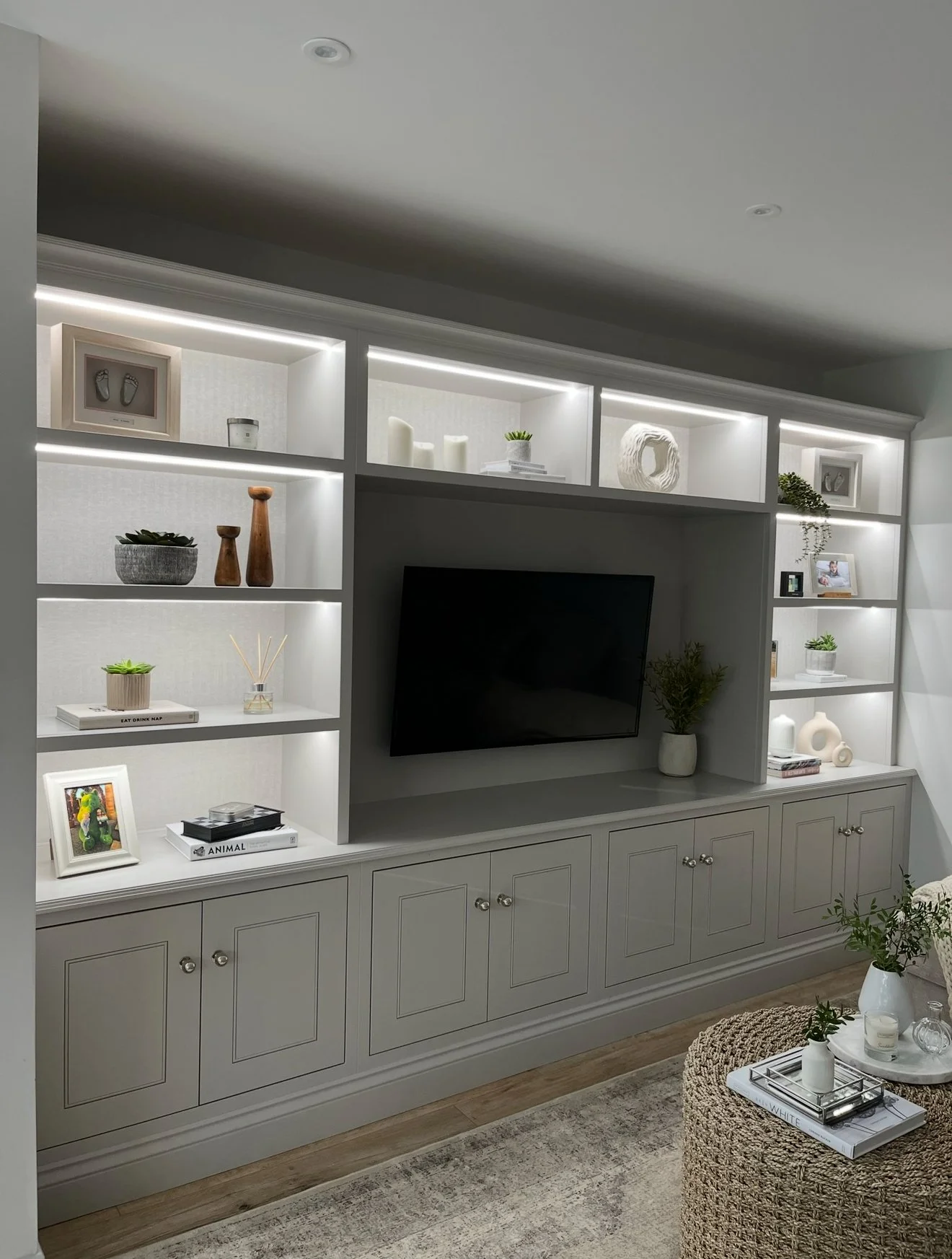 Living room wall unit with shelves and cabinets, decorated with books, vases, candles, framed photos, and plants, with a flat-screen TV in the center.