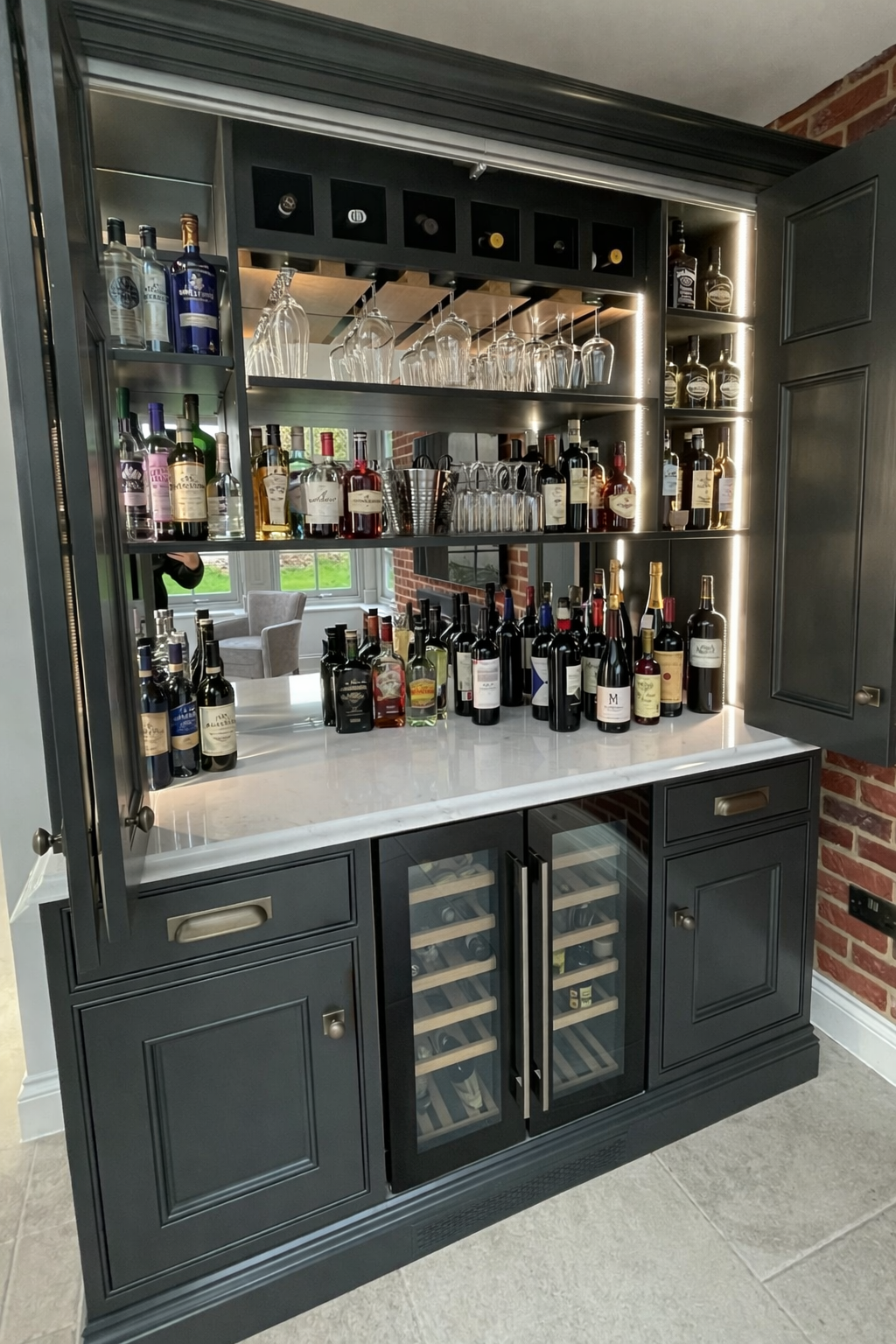 A dark gray built-in bar cabinet with a marble countertop, containing various bottles of wine and liquor, glassware, and a wine cooler with bottles inside. A window with greenery outside is reflected in the upper mirror panel.