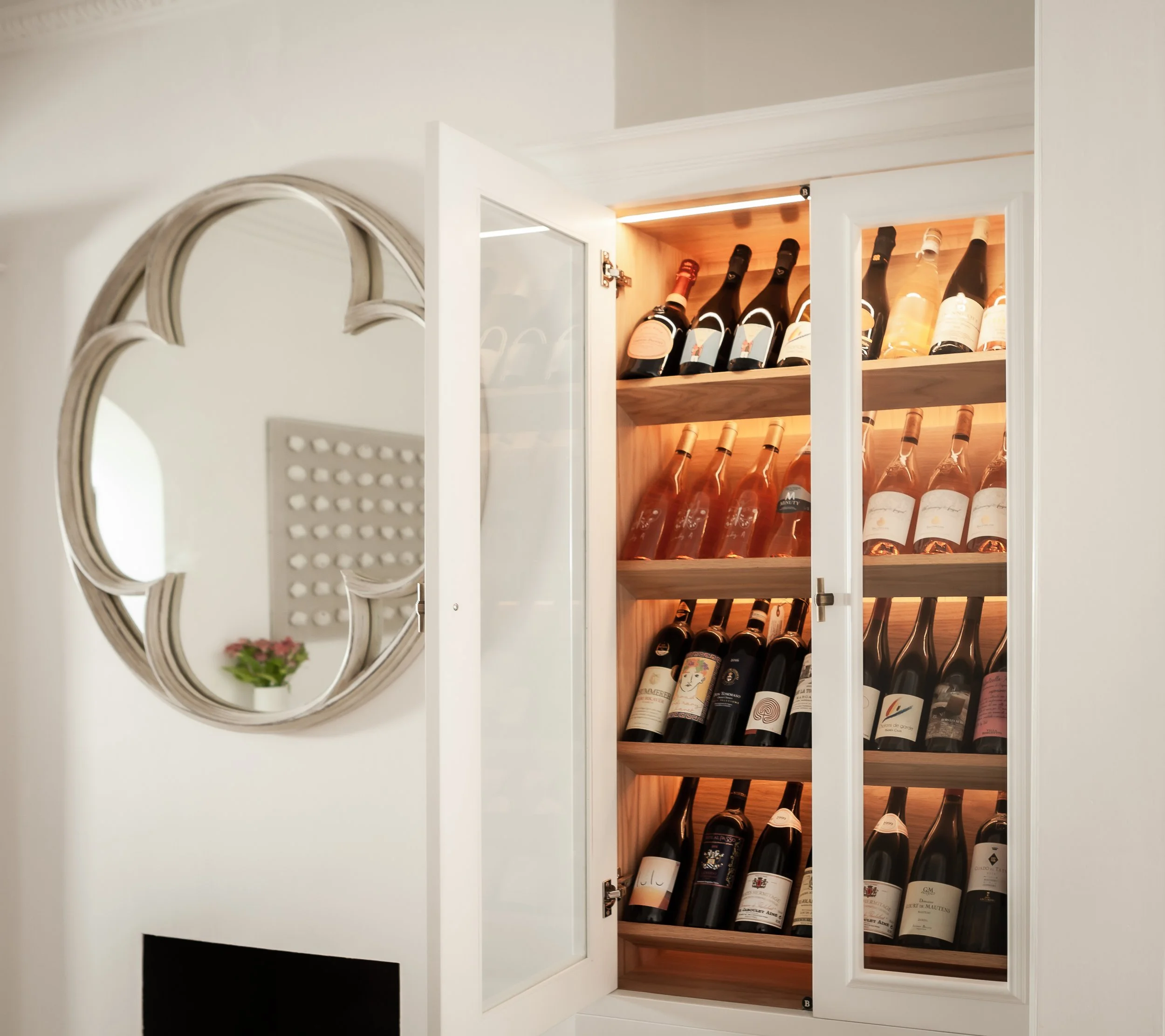 A glass-front cabinet with shelves filled with various bottles of wine, illuminated from behind, in a room with white walls and a decorative mirror.