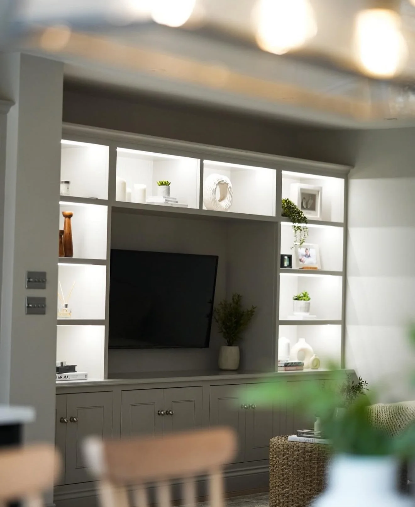 Living room with a wall-mounted TV, white shelving unit with decorative items, plants, and framed photos.