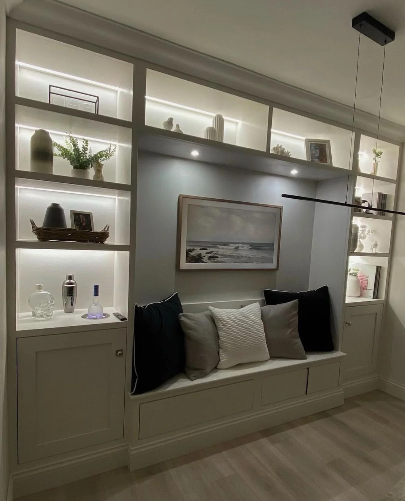 A built-in wall storage unit with open shelves illuminated by lighting, containing decorative objects like vases, framed photos, and small sculptures. Below the shelves is a sitting area with a bench, adorned with several pillows, and a framed landsc