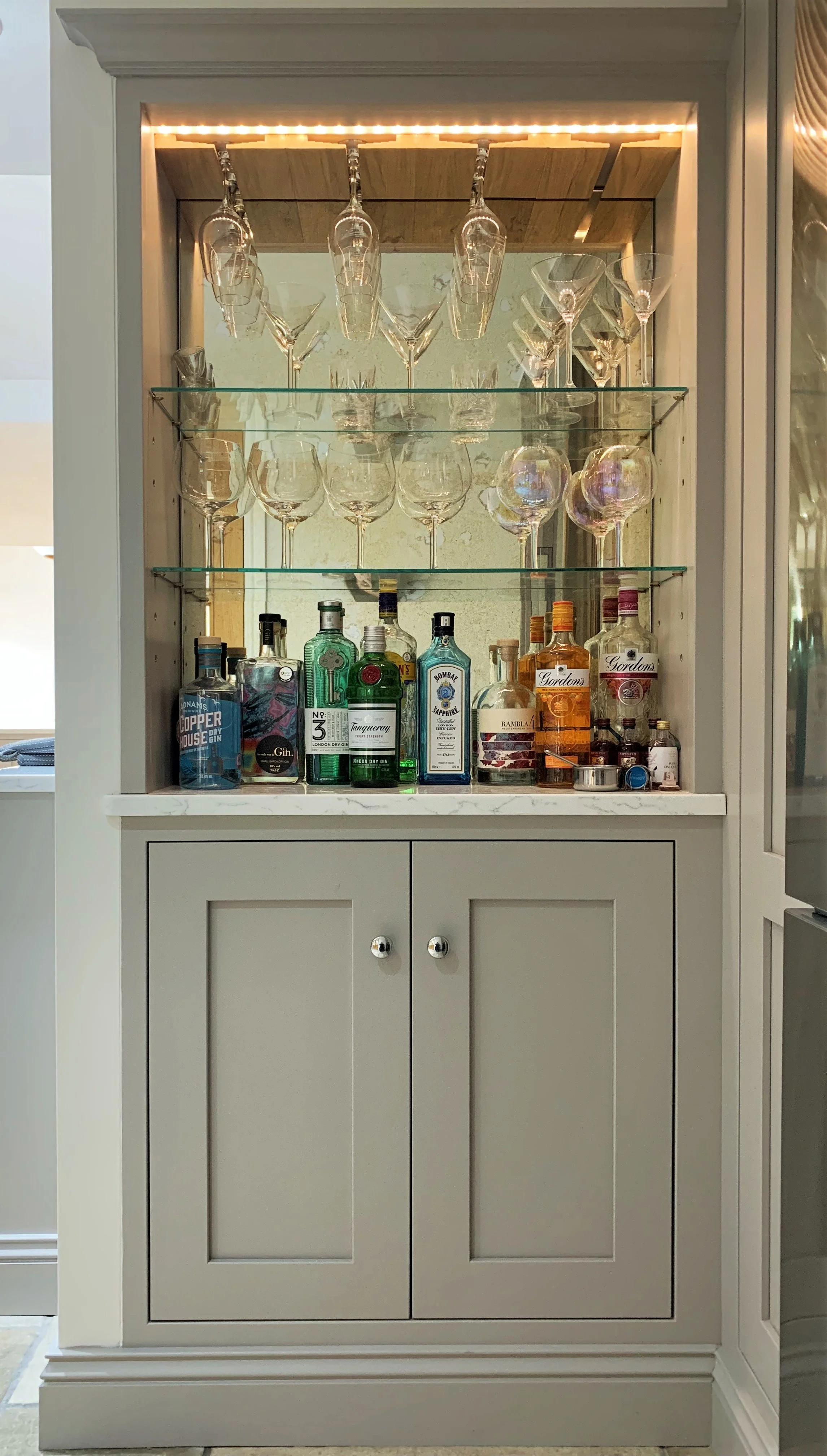 A home bar cabinet with glasses hanging upside down on a rack and bottles of alcohol on the countertop, beneath glass shelves.