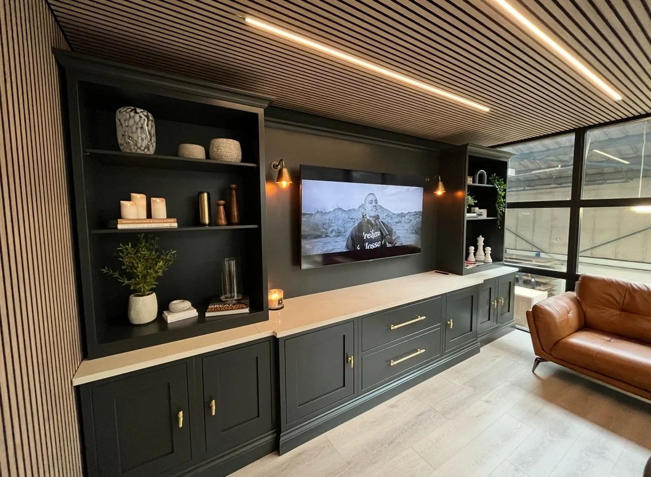 Modern living room featuring a black built-in media center with open shelves decorated with vases, candles, books, and decor, a large flat-screen TV, a brown leather sofa, and floor-to-ceiling windows with grid panes showing an outdoor view.