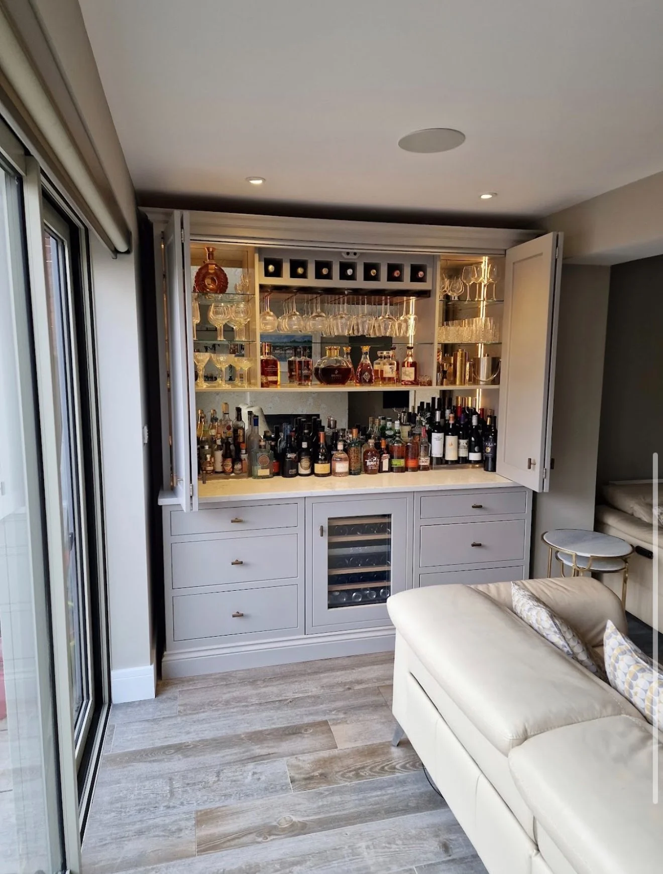 Home bar with open cabinets filled with various liquor bottles, glasses, and wine bottles, adjacent to sliding glass doors and a cream sofa with patterned pillows.