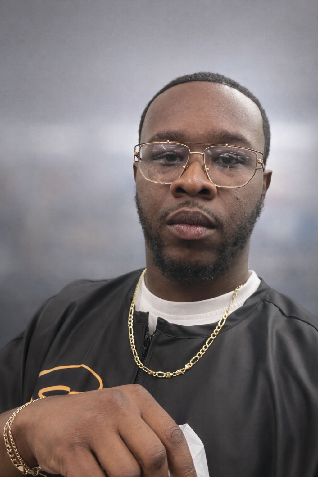 A man with glasses, a beard, and jewelry, wearing a black jacket and a white shirt, holding a white object in his hand, standing indoors near a wall with a framed picture.