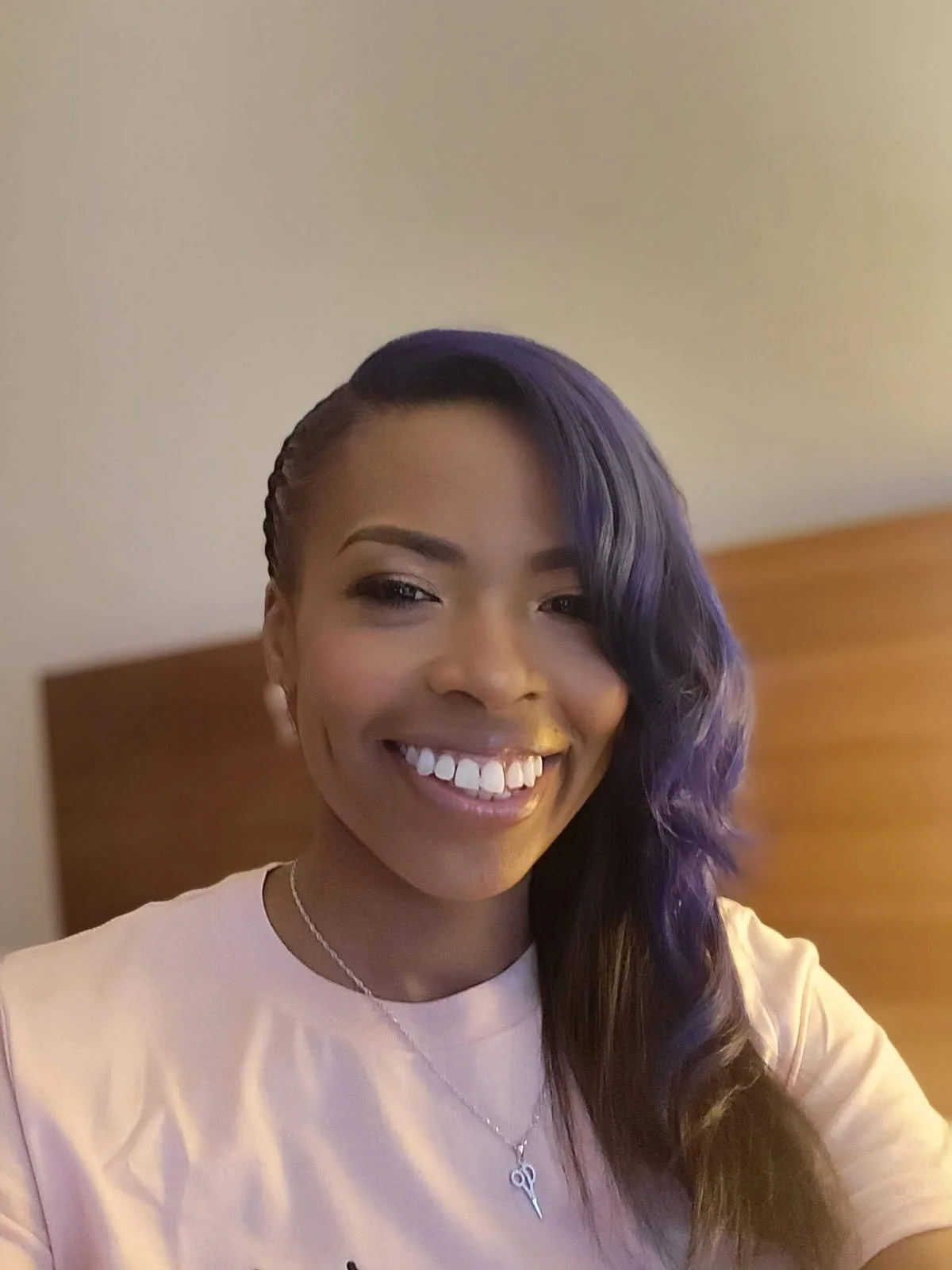 A smiling woman with dark purple hair styled to the side, wearing a light pink shirt, a silver necklace, and earrings, sitting in front of a beige wall and wooden headboard.
