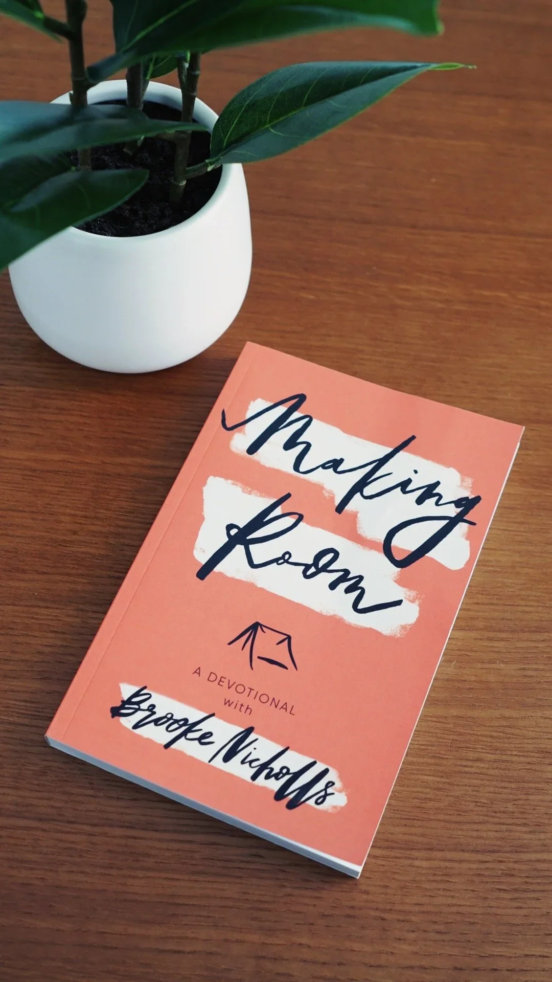 A pink devotional book titled 'Making Room' by Brooke Nicholson resting on a wooden surface next to a potted plant with dark green leaves.