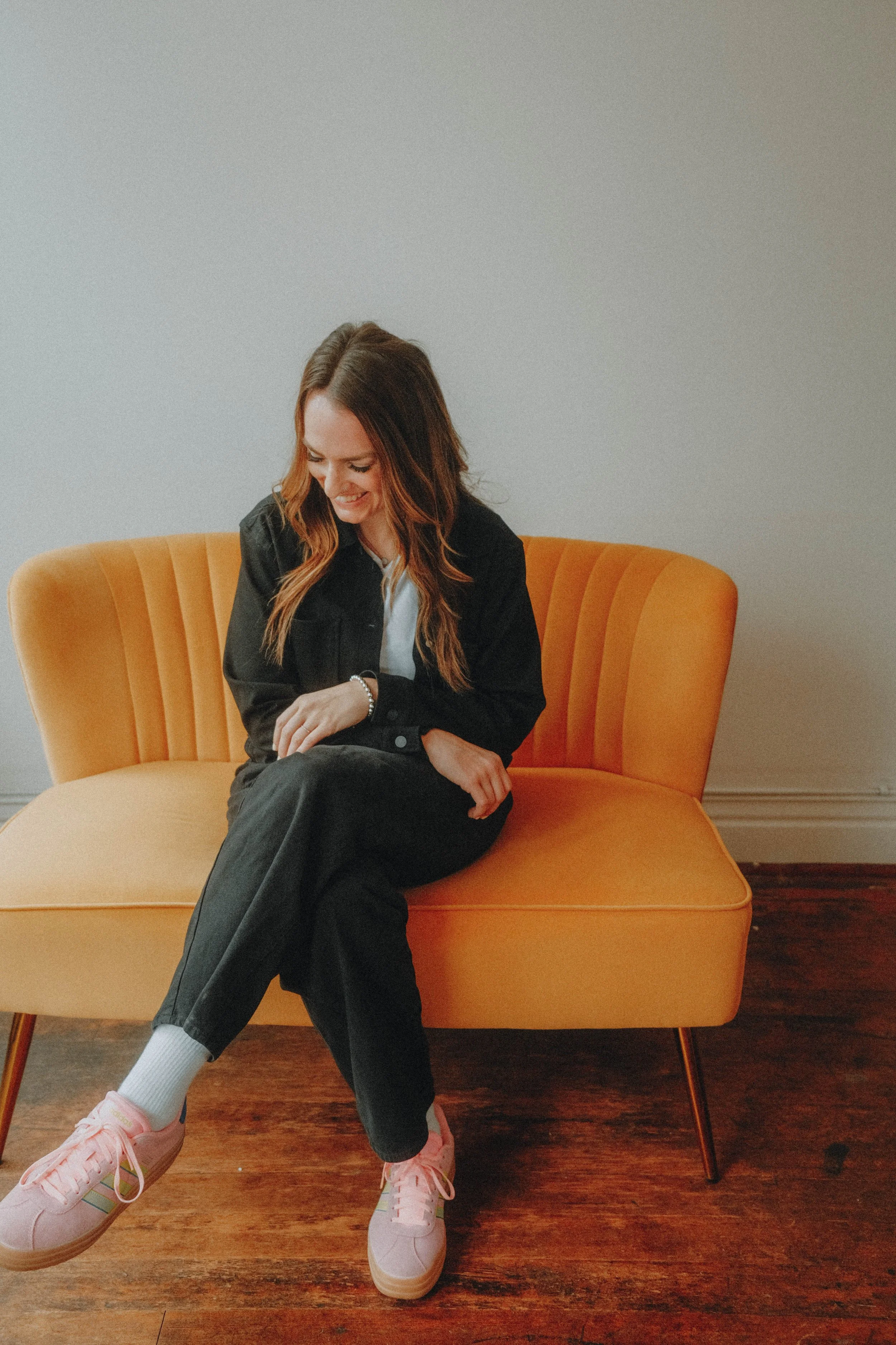 A young woman with long brown hair, wearing a black jacket, white shirt, black pants, white socks, and pink sneakers, sitting on an orange mid-century style chair against a plain beige wall, smiling and looking down.