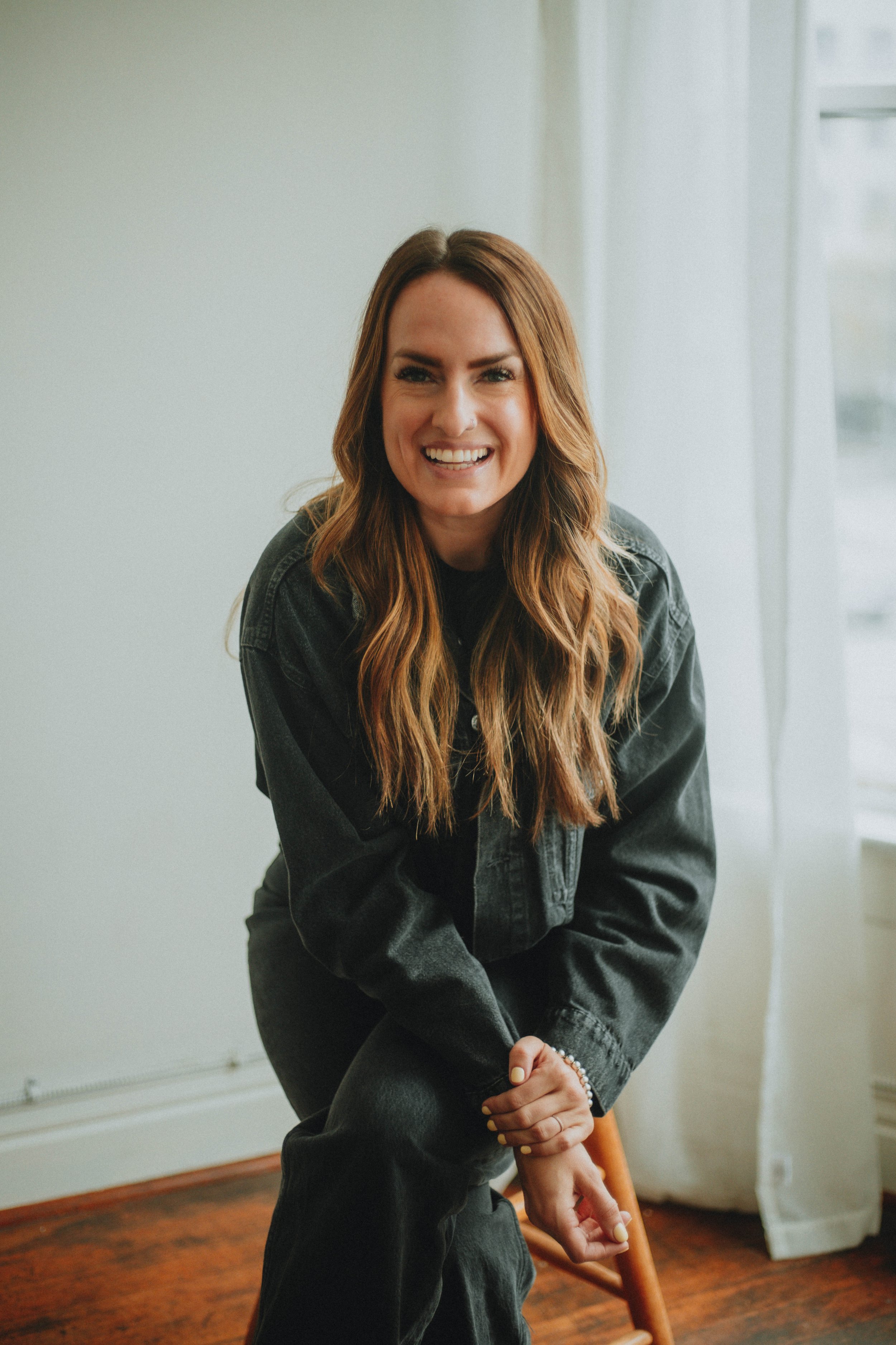 A woman with long, wavy, reddish-brown hair smiling and sitting in a room with white walls and wooden floors, wearing a black denim jacket.