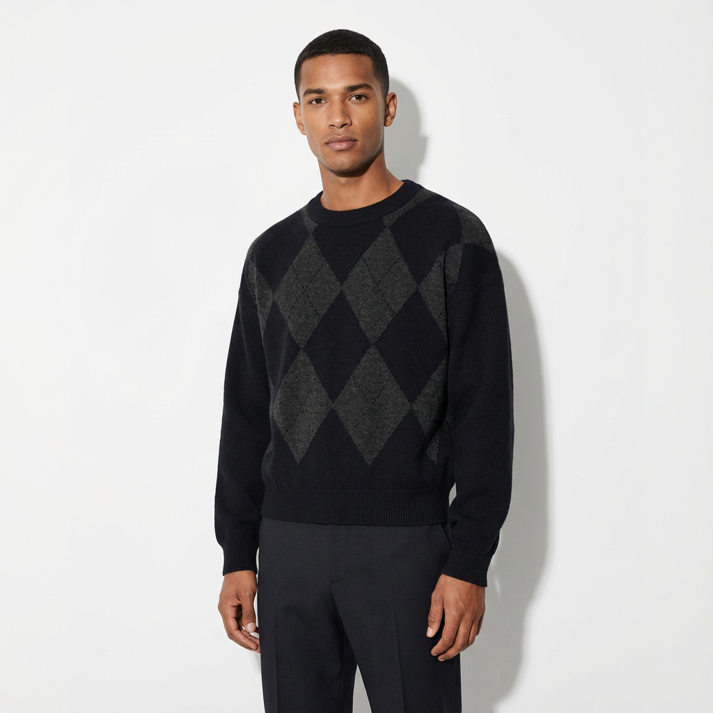 Man standing against a white wall, wearing a black sweater with an argyle pattern and black pants.