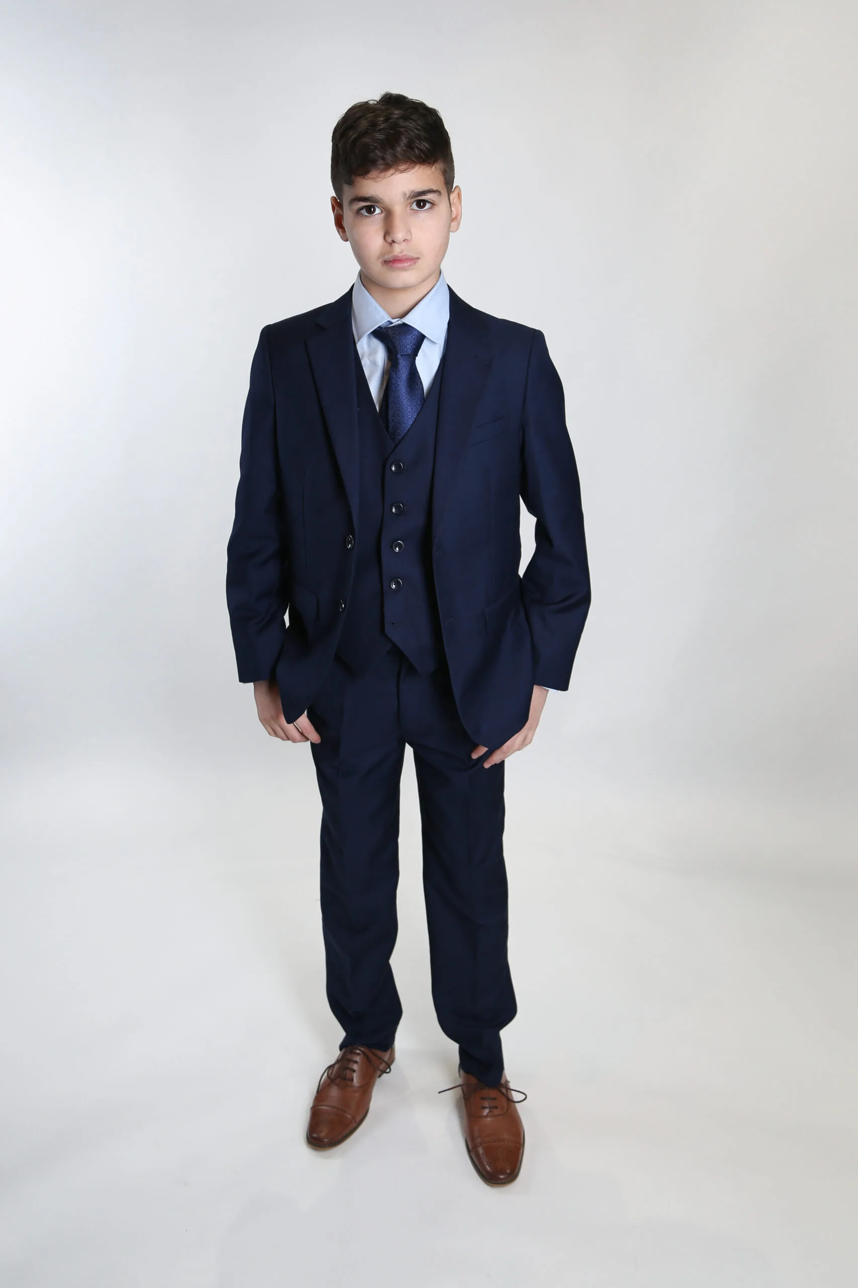 A young boy in a navy blue suit, white shirt, and blue tie, standing against a plain white background.