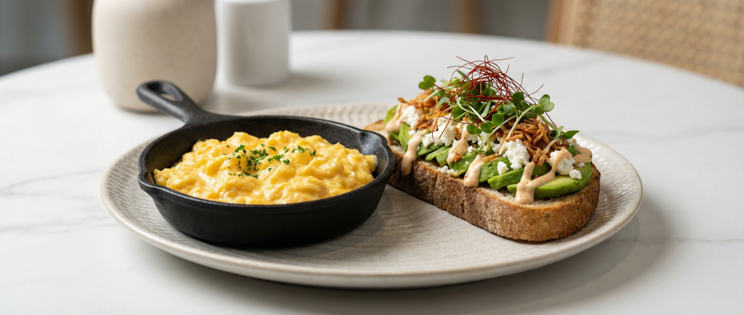 A plate with scrambled eggs in a black skillet and an avocado toast topped with microgreens, shredded chicken, crumbled cheese, and drizzled with sauce on a white textured plate.