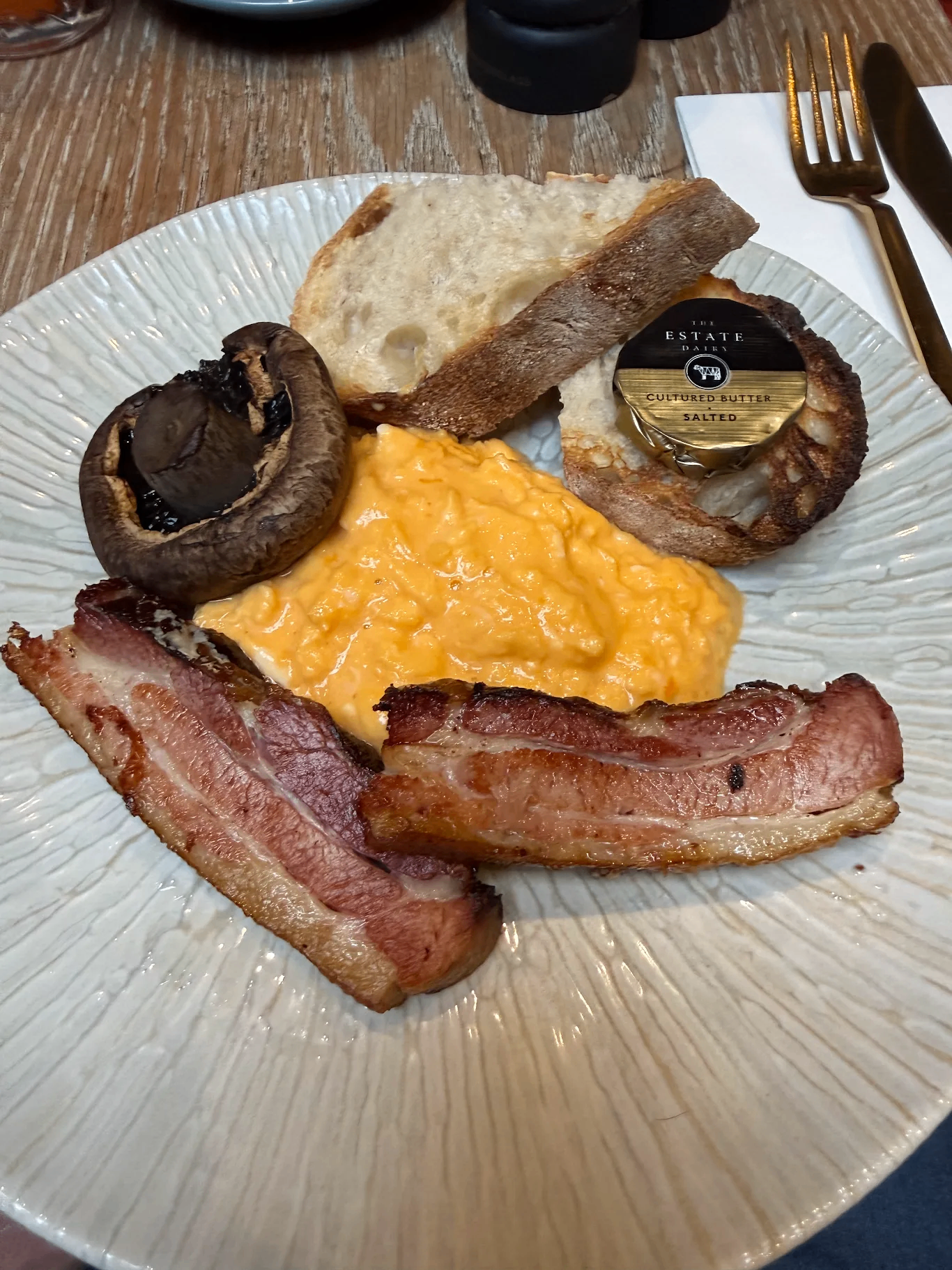 Plate of breakfast food with scrambled eggs, two strips of bacon, toast, a grilled mushroom, and a baked onion with a packet of cultured salted butter.
