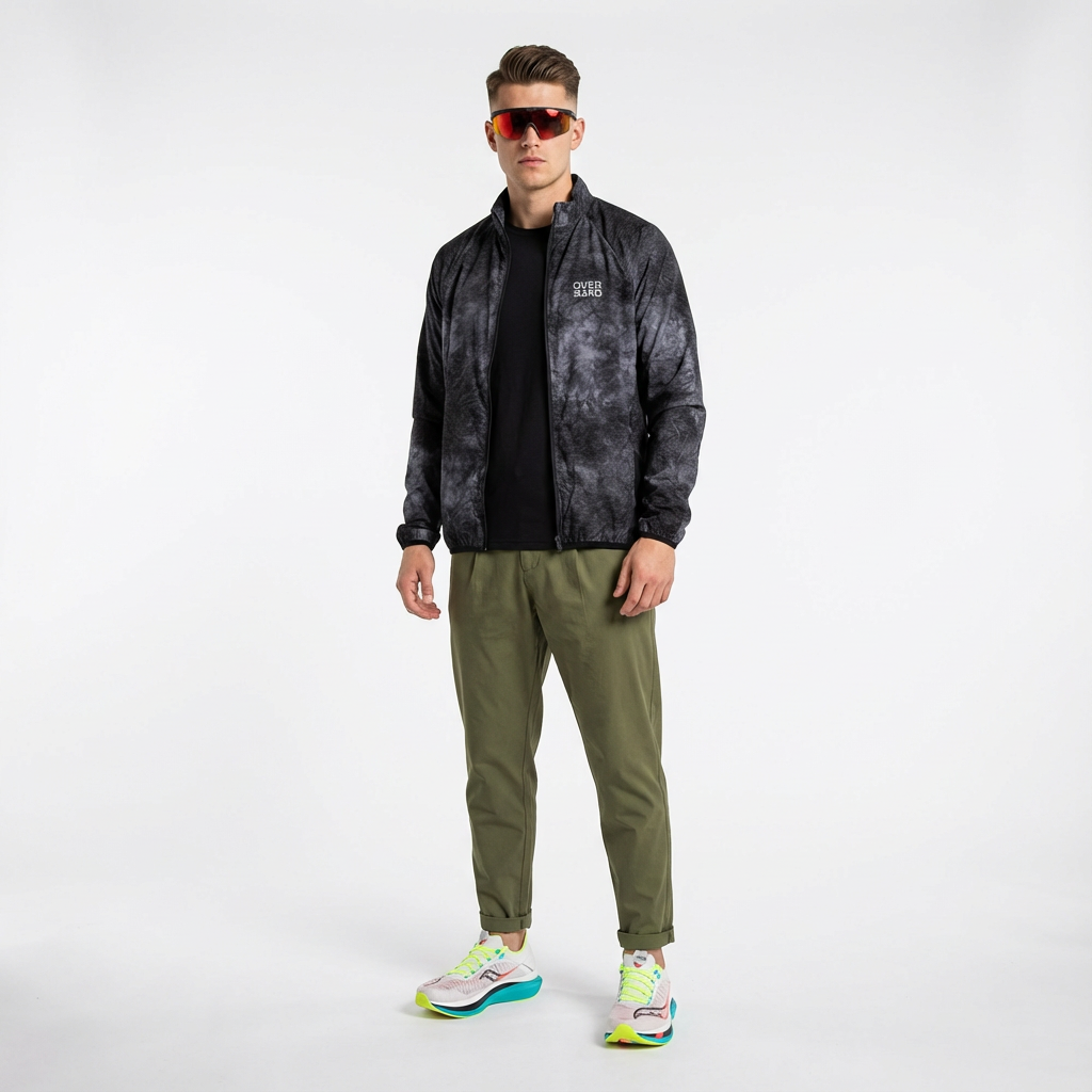 A young man wearing a black and grey patterned zip-up jacket, black shirt, olive green pants, colorful running shoes, and sporty sunglasses standing against a plain white background.