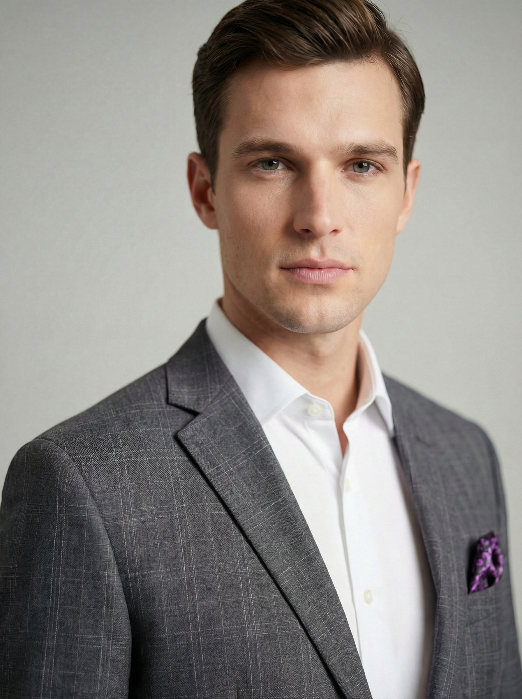Portrait of a young man with fair skin, brown hair, blue eyes, wearing a gray checkered suit jacket over a white dress shirt and a purple pocket square, against a plain background.