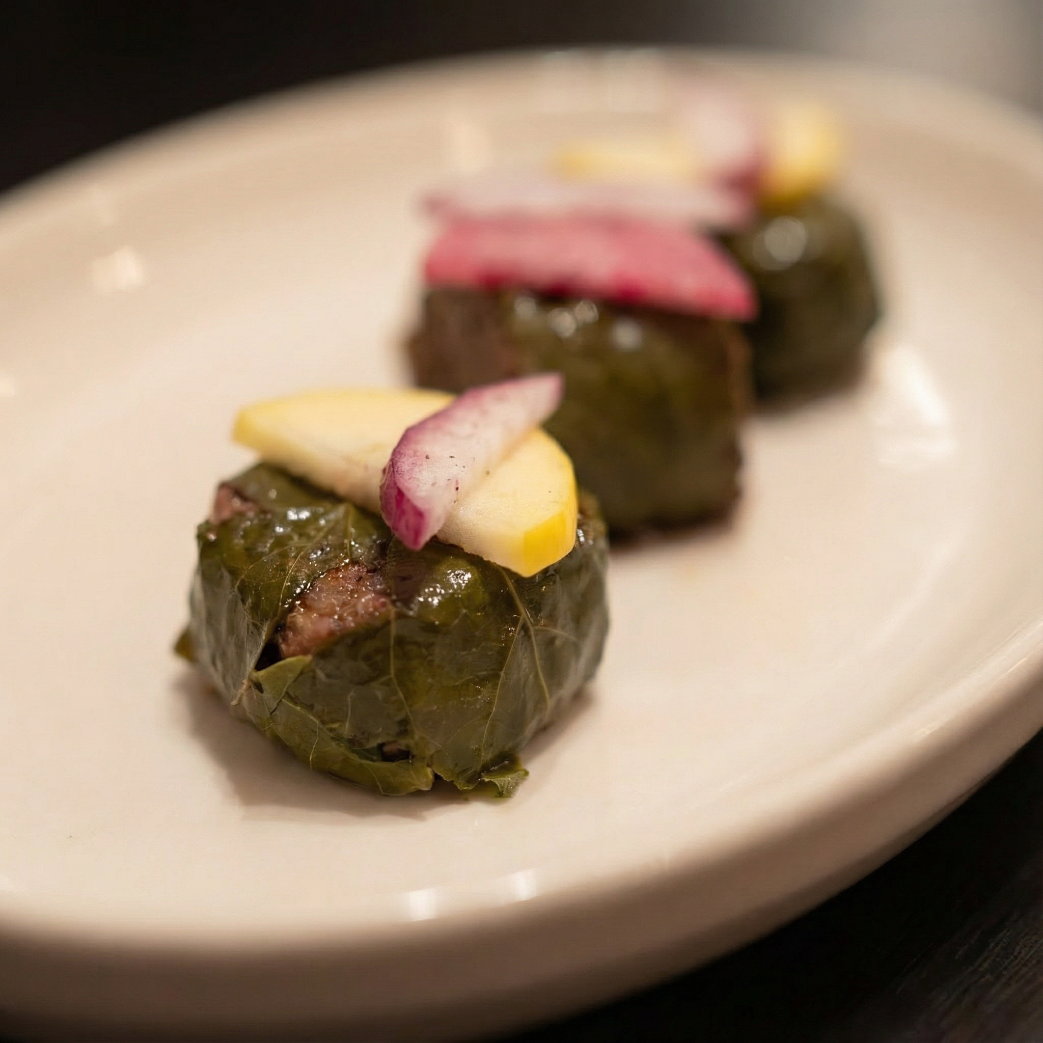 Three pieces of stuffed grape leaves on a white oval plate, each topped with slices of radish and pickled vegetables.