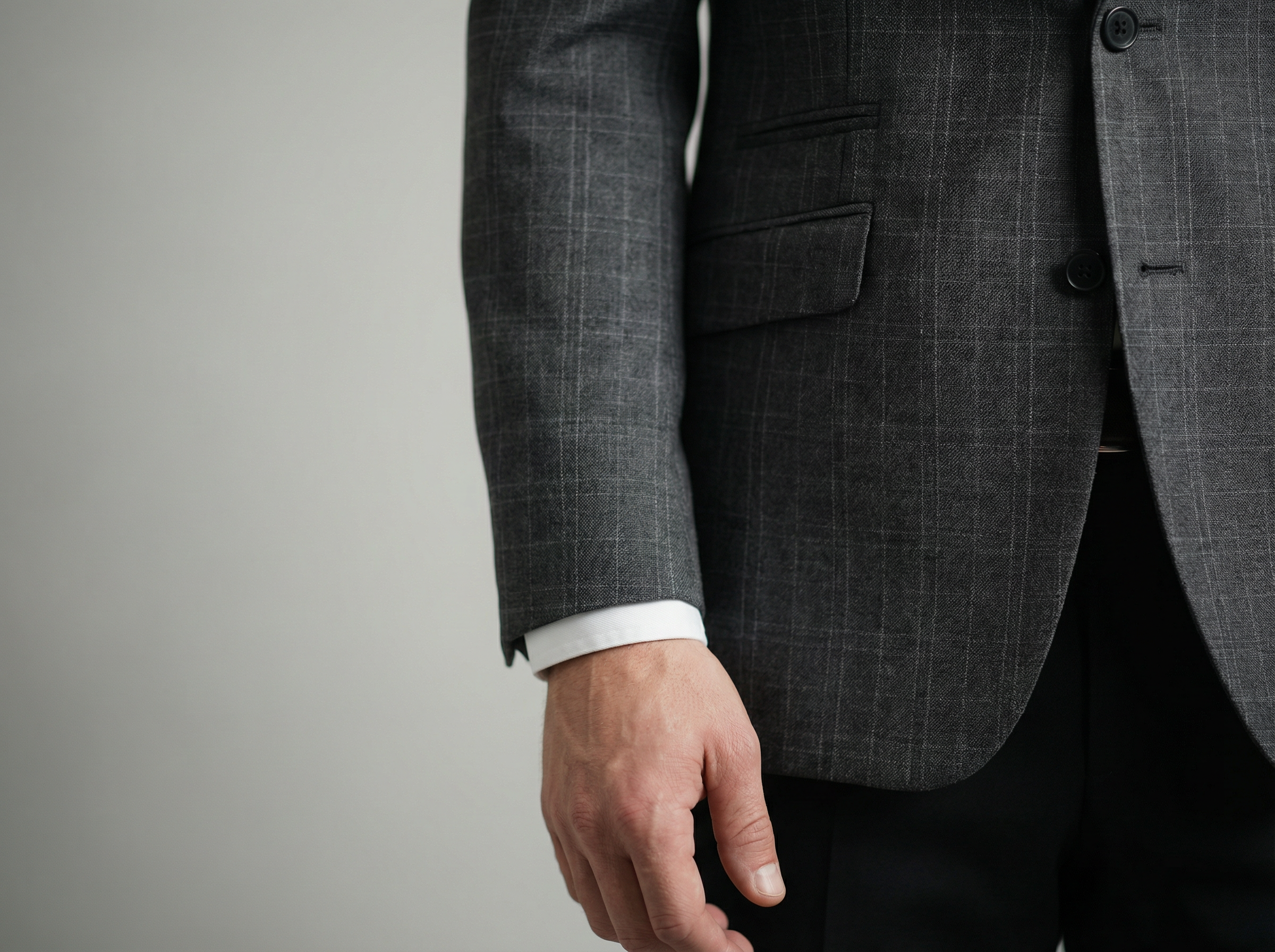 Close-up of a man in a dark gray suit with a subtle checkered pattern, white shirt sleeve visible, standing against a plain background.