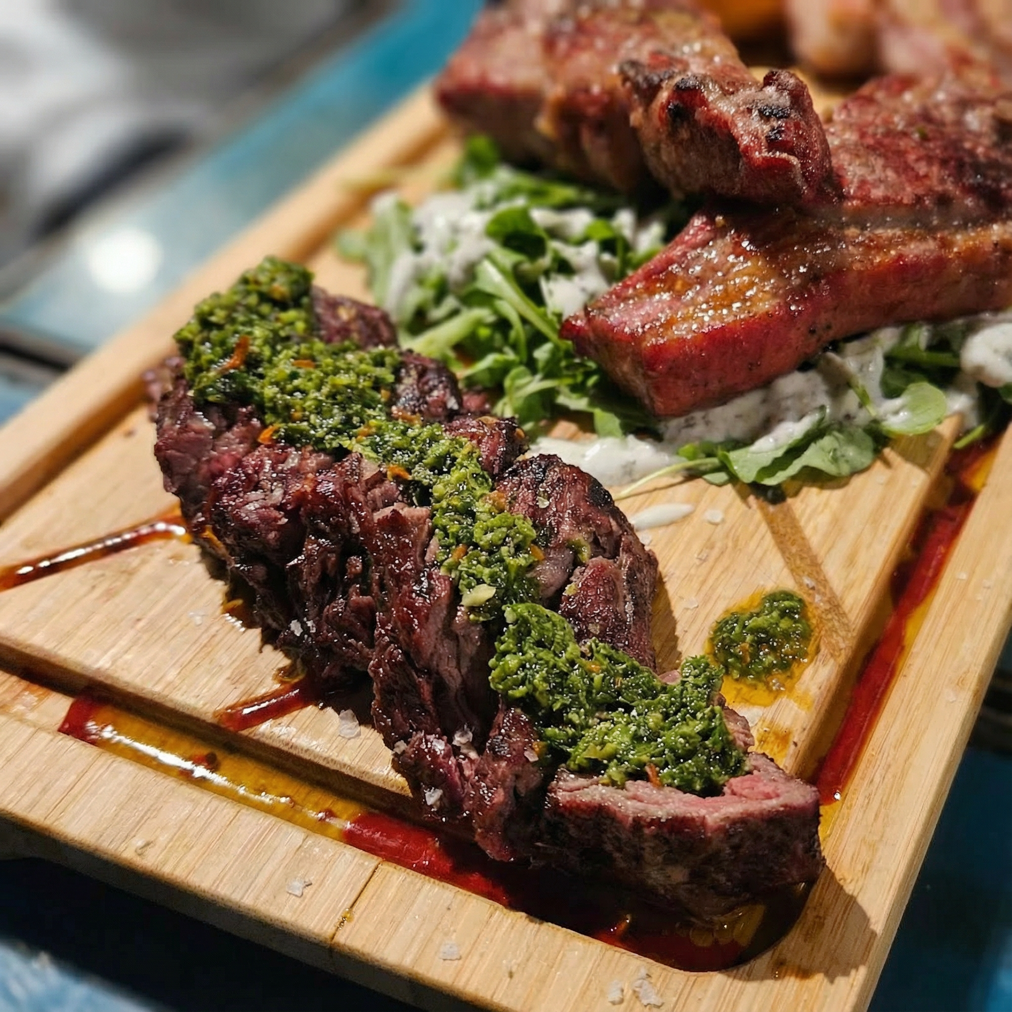 Close-up of a wooden platter with slices of steak topped with chimichurri sauce, grilled beef ribs, and a side salad with creamy dressing.