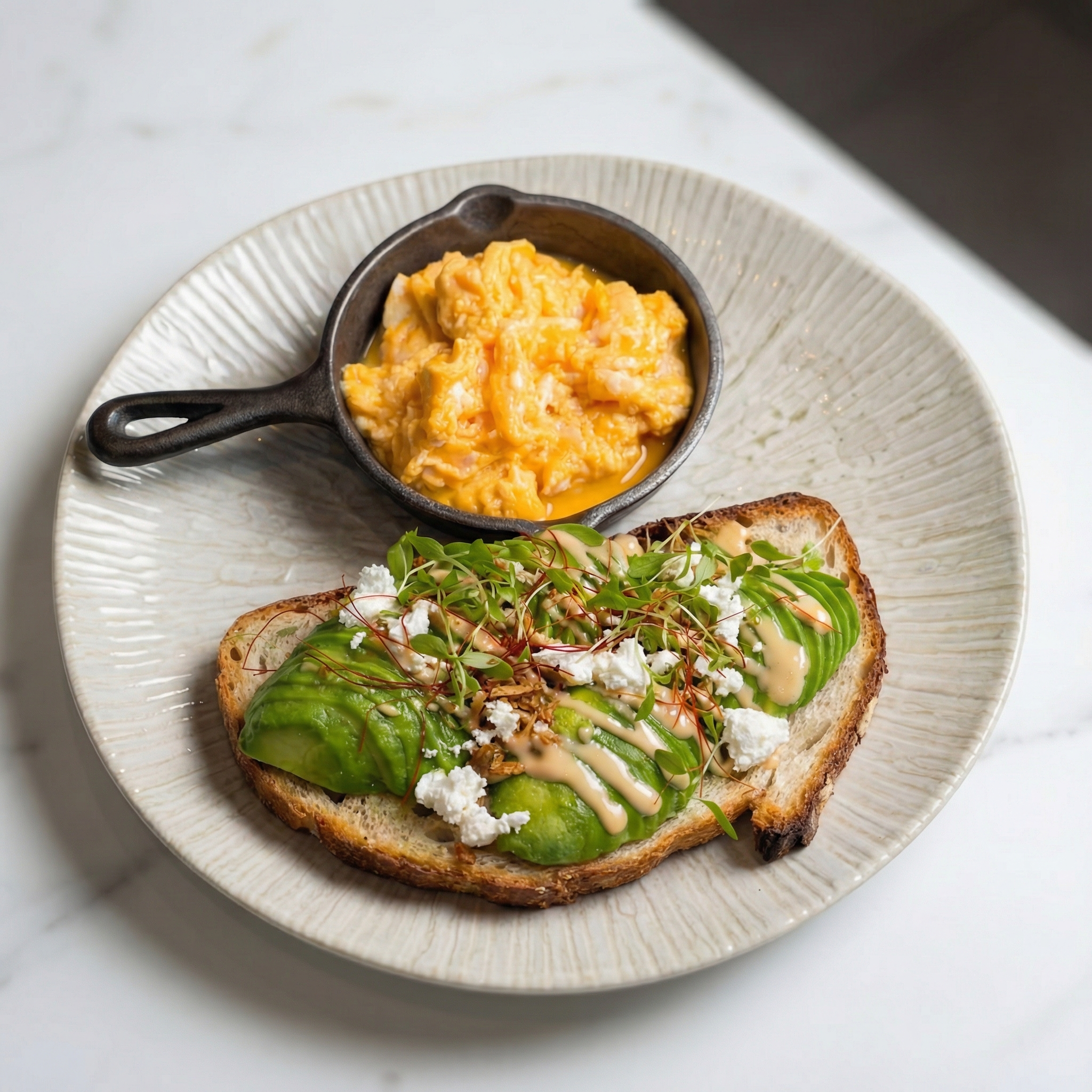 Scrambled eggs in a cast iron skillet, avocado toast with microgreens and drizzled sauce on a white plate.