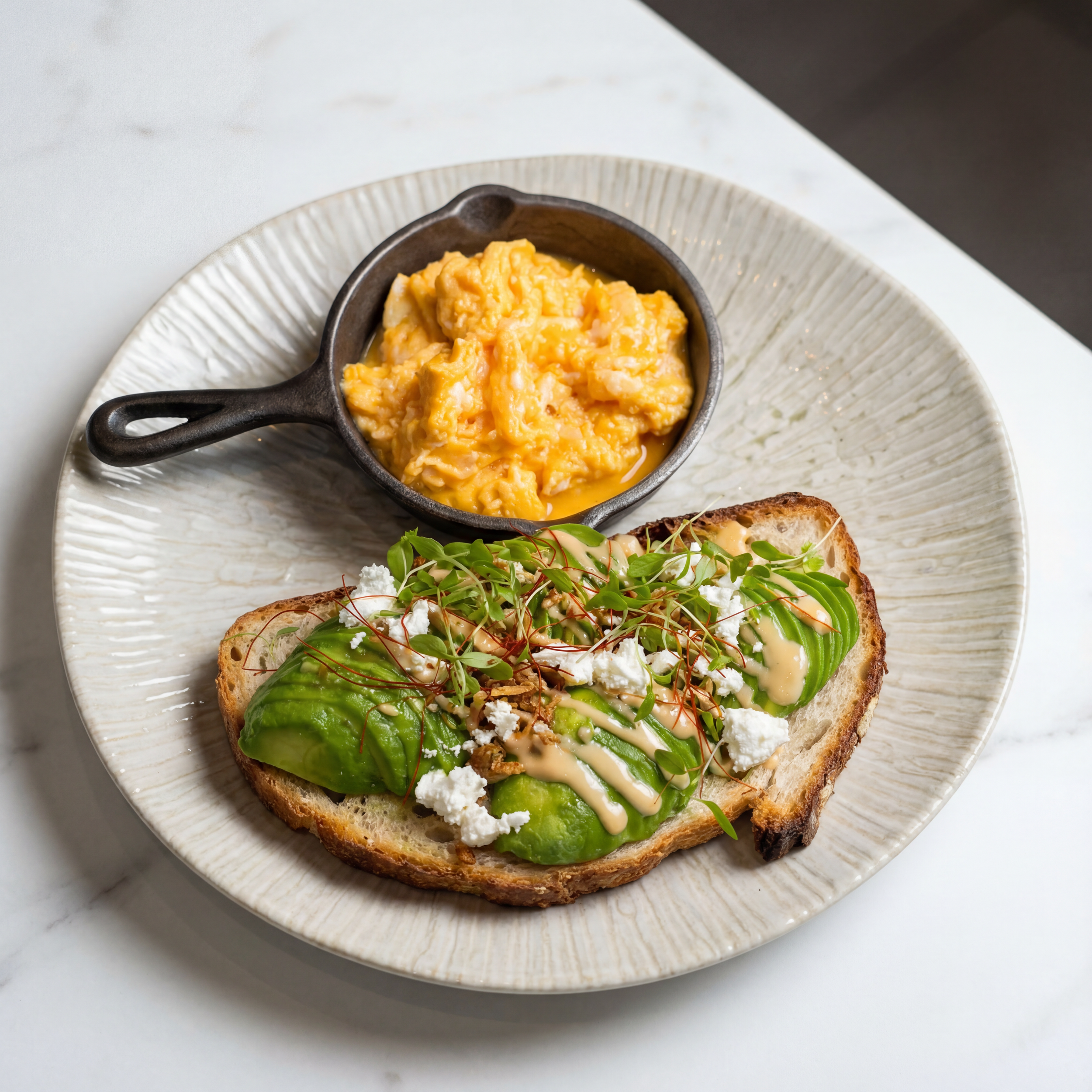 Scrambled eggs in a small cast-iron skillet and toasted bread with avocado, microgreens, crumbled cheese, and drizzled sauce on a white plate