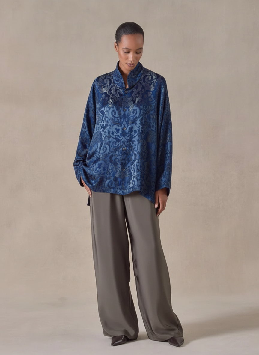 A woman in a shiny blue patterned button-up shirt and loose gray pants standing against a plain background.