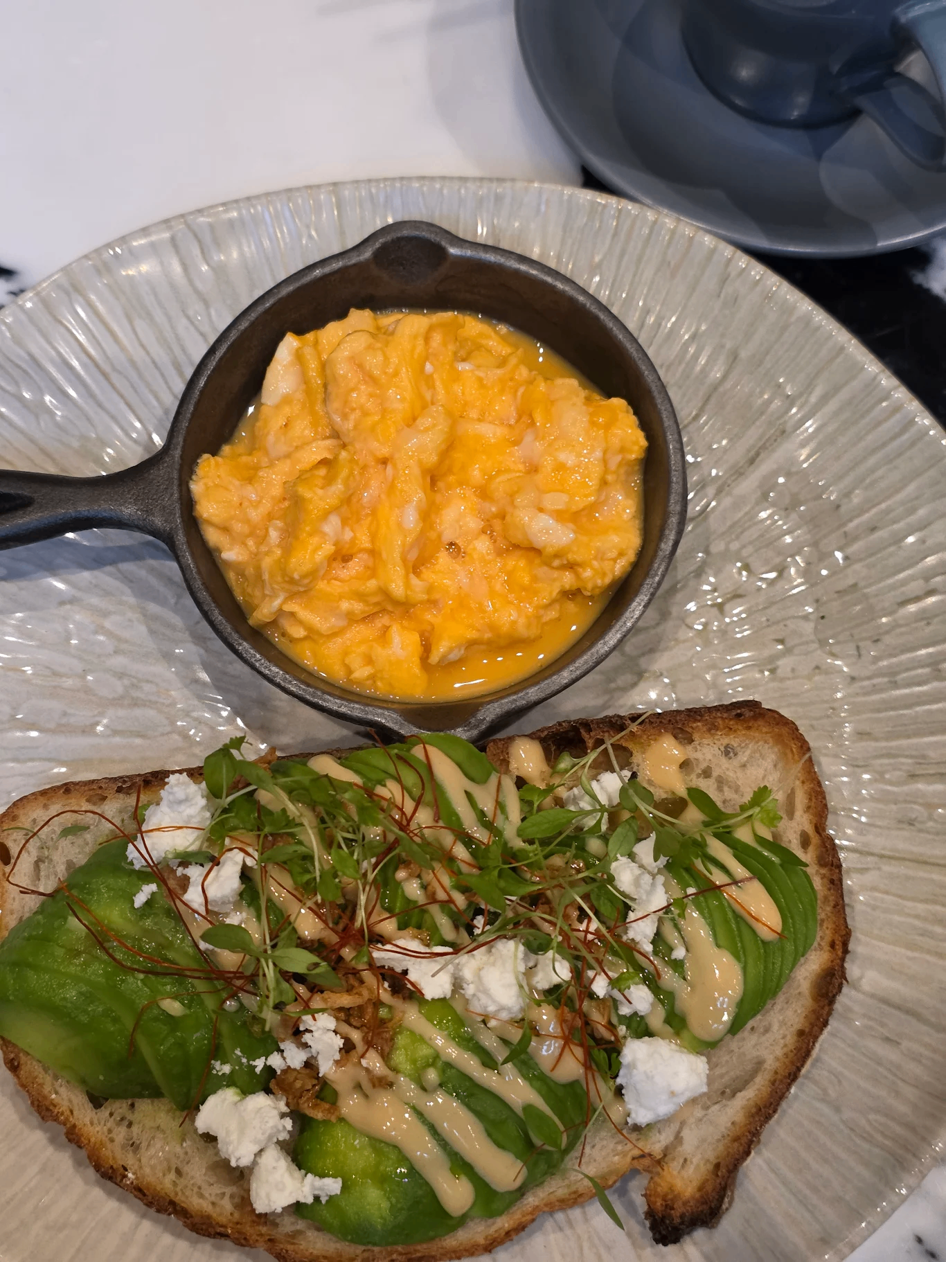 Scrambled eggs in a small cast iron skillet and avocado toast with microgreens, crumbled cheese, and drizzle of sauce on a decorative plate.