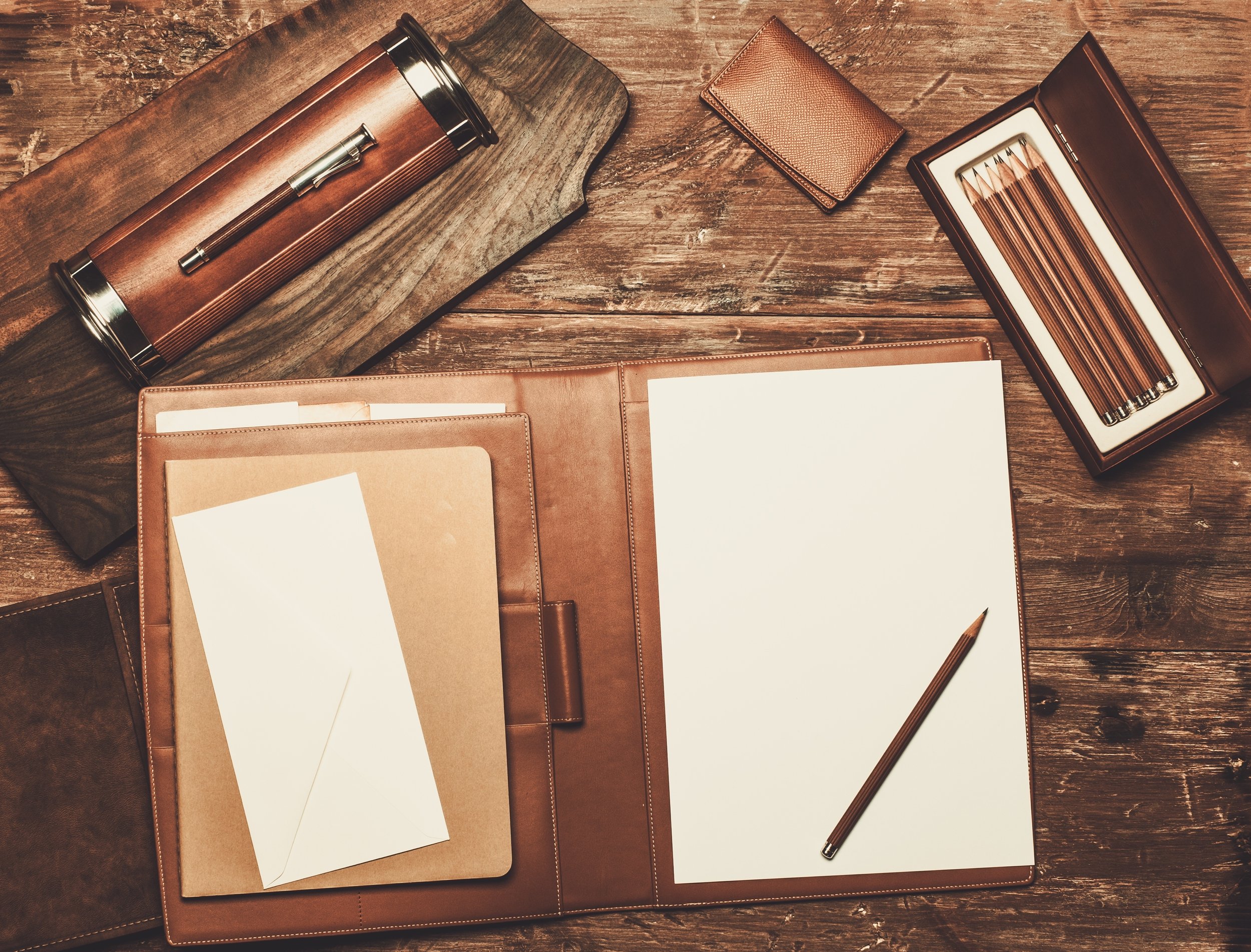 A wooden desk with various leather accessories, including a brown leather portfolio with papers, a small envelope, a leather-wrapped thermos, a box of colored pencils, and a leather wallet, all arranged on the surface.