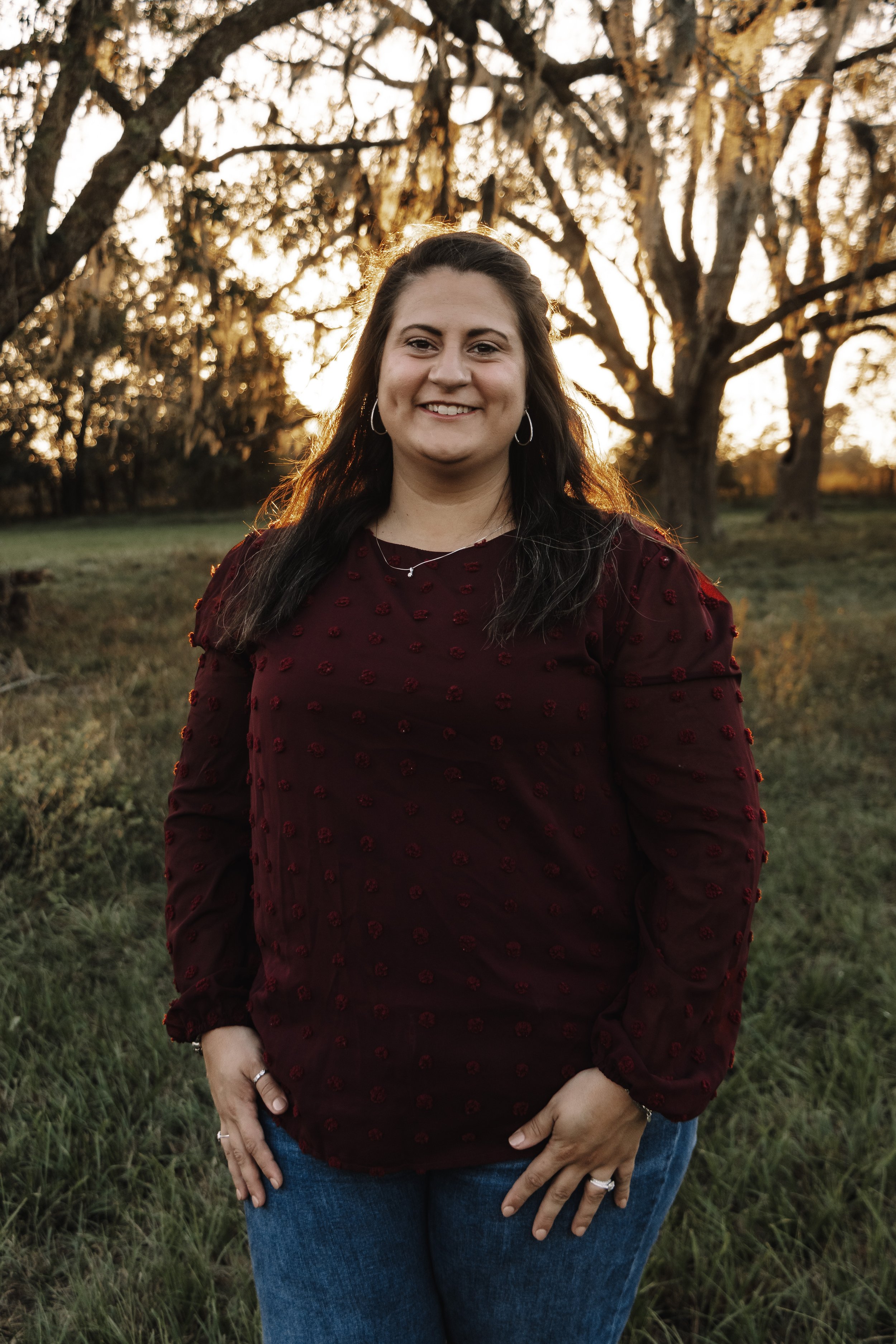 A woman with long dark hair, wearing a maroon top with textured details and blue jeans, stands outdoors at sunset with trees in the background.