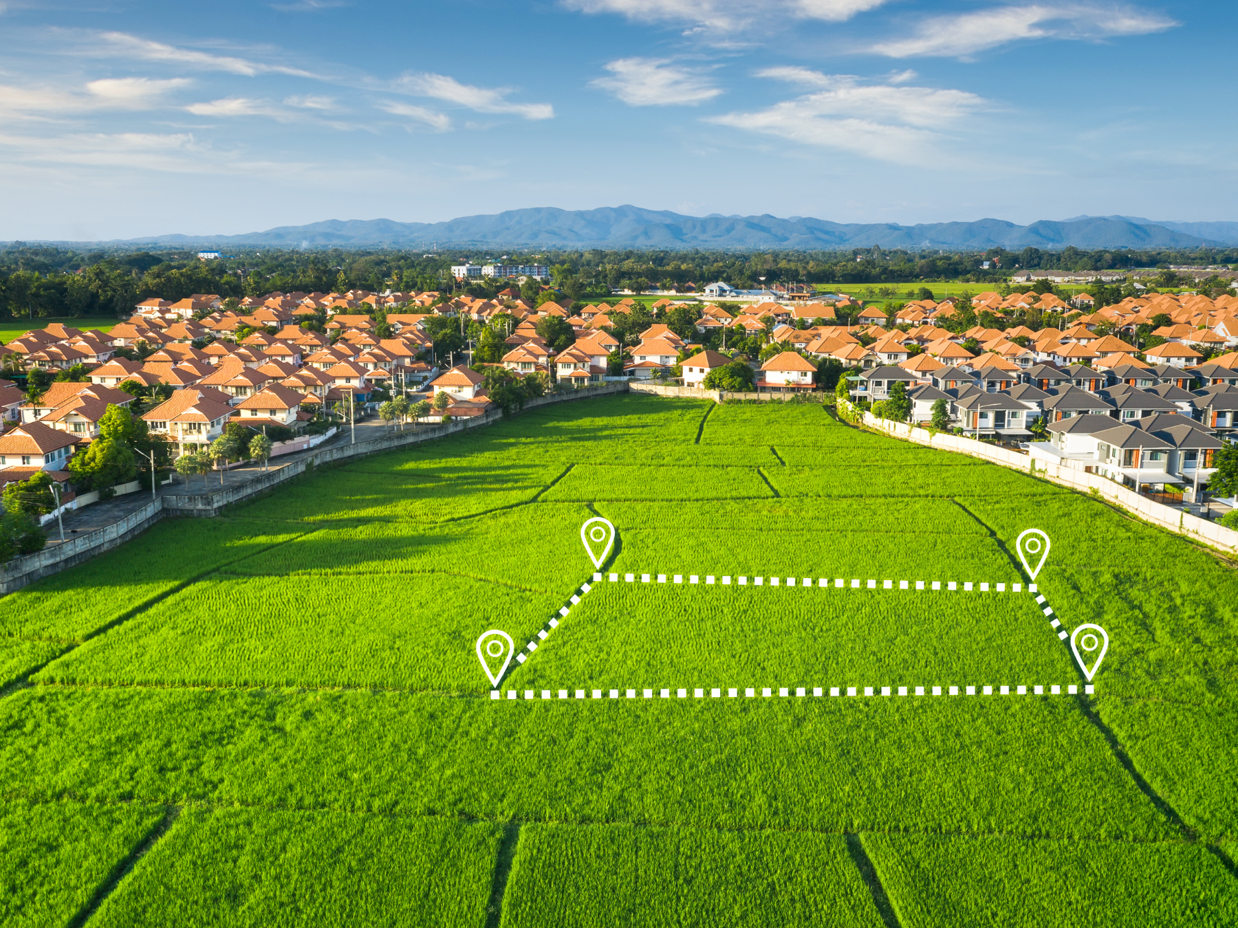 Aerial view of a green rice field with a dotted line demarcation and four location markers. Surrounding the field are residential houses with orange and gray roofs, with mountains in the background and a partly cloudy sky.