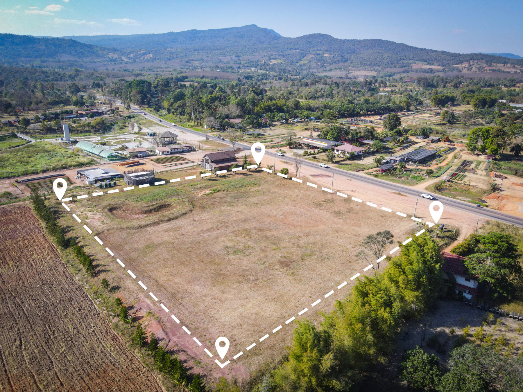 Aerial view of a large empty plot of land bordered by a hedge, with a few trees and buildings on the edges. The land is next to a road with several cars and is surrounded by a mix of rural and residential areas, with mountains in the background.