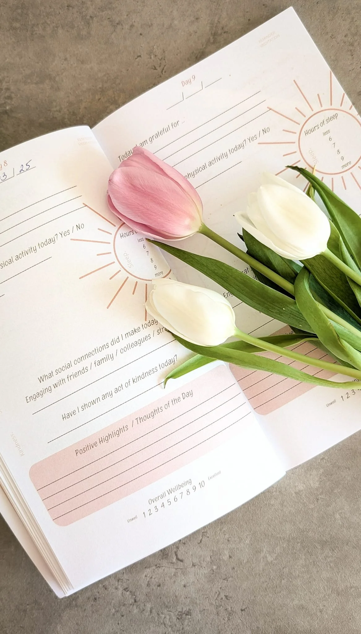 Open journal with a prompt to reflect on the day, including questions about social interactions, acts of kindness, and positive highlights. There are three tulips—two white and one pink—resting on the journal pages.