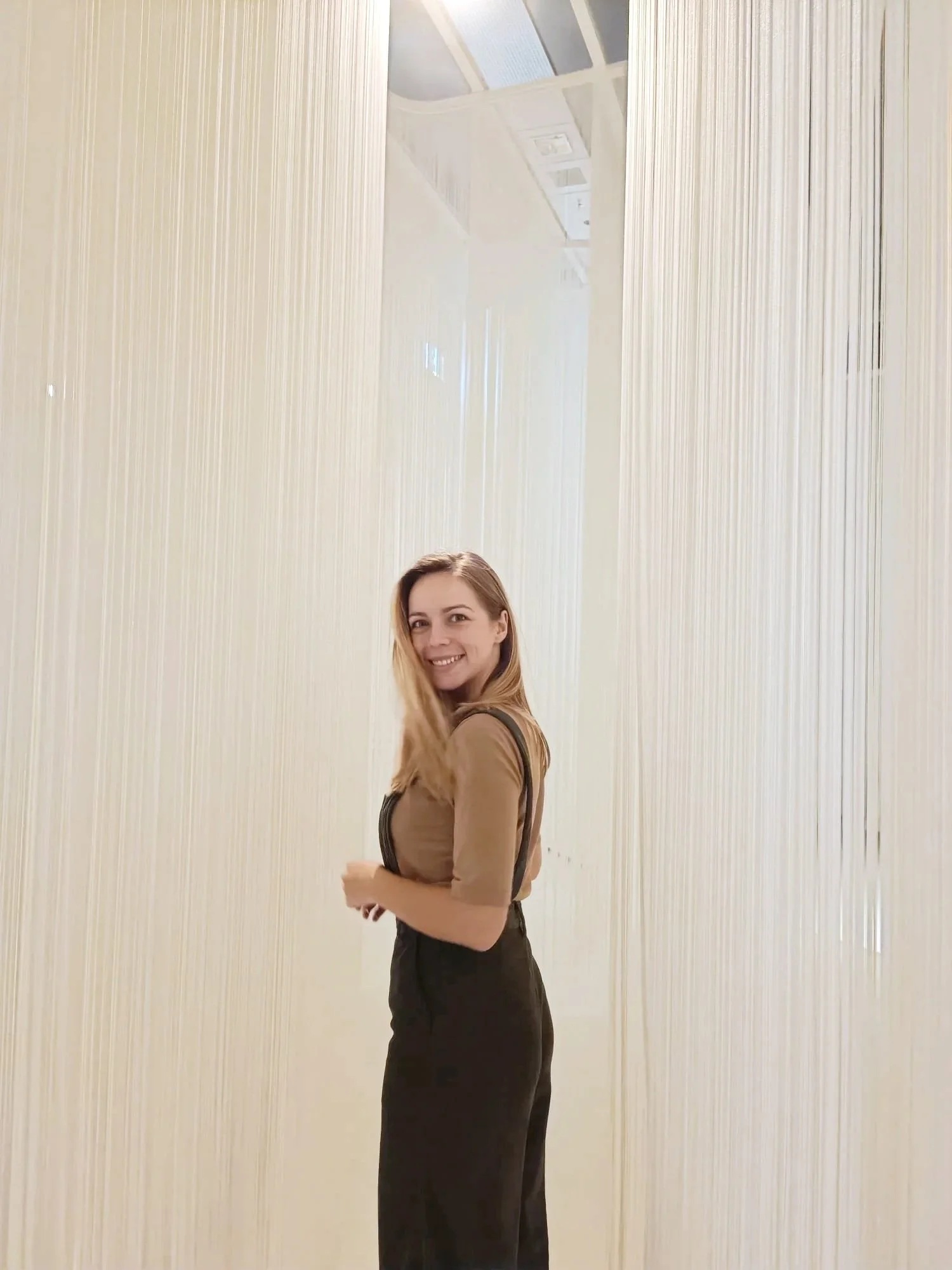 Woman with long blonde hair smiling and looking at camera, standing in front of a vertical, light-colored textured wall, wearing a beige top and black pants.