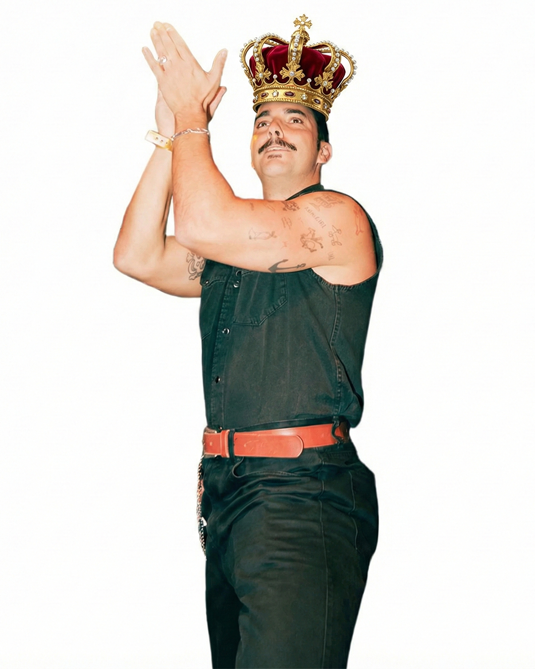 Hombre con corona real, peinado oscuro, barba y tatuajes en el brazo, vestido con camiseta sin mangas negra y cinturón naranja, posando con las manos juntas en forma de oración, en fondo blanco.