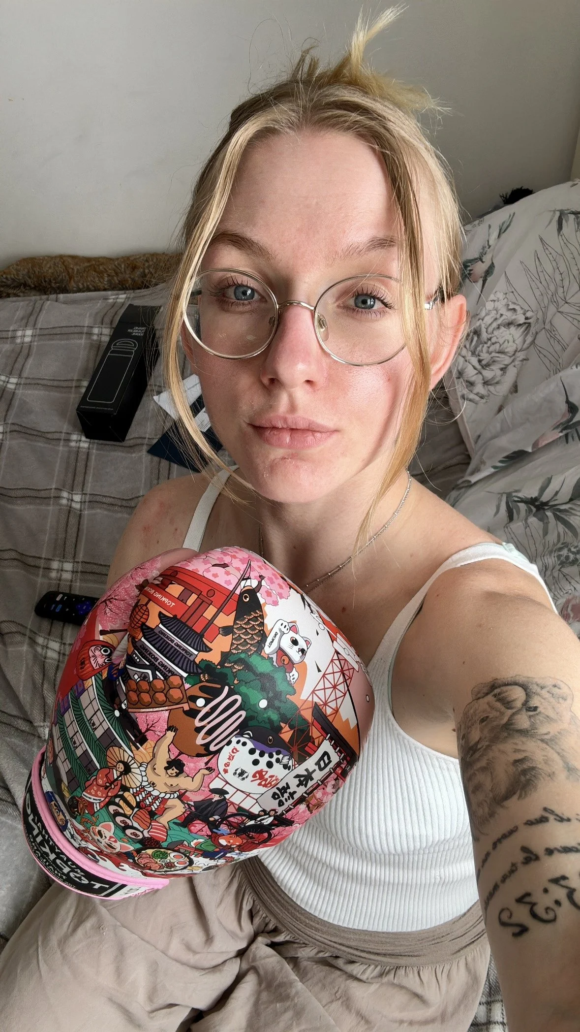 A young woman with blonde hair, glasses, and tattoos on her arm, wearing a boxing glove with Japanese-themed illustrations.