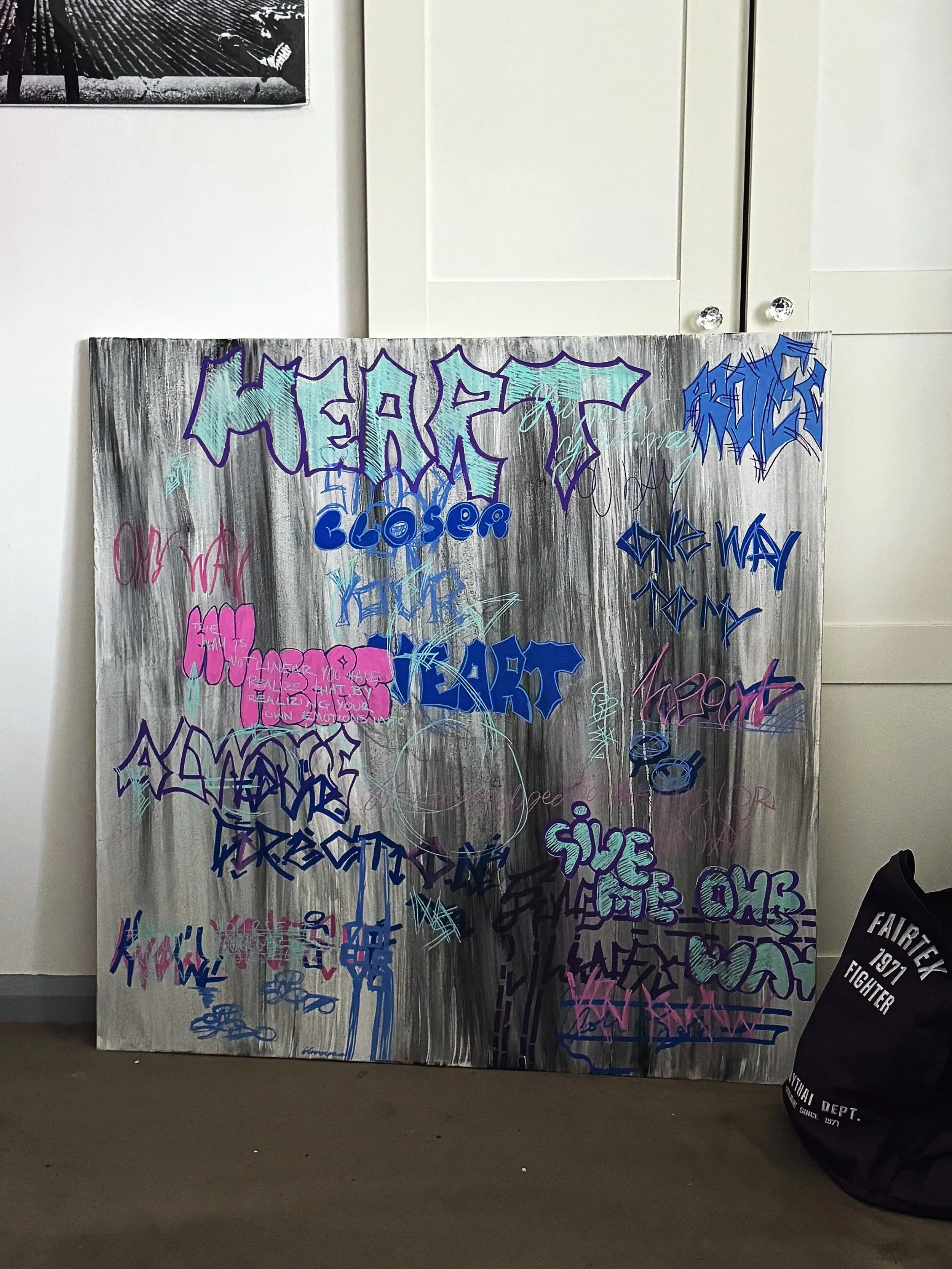 Colorful graffiti art on a wooden board with words like "HERE," "CLOSER," "ONE WAY TO MY", and "HEART", written in various styles and colors, mostly purple, blue, pink, and teal, with a gray and black painted background.